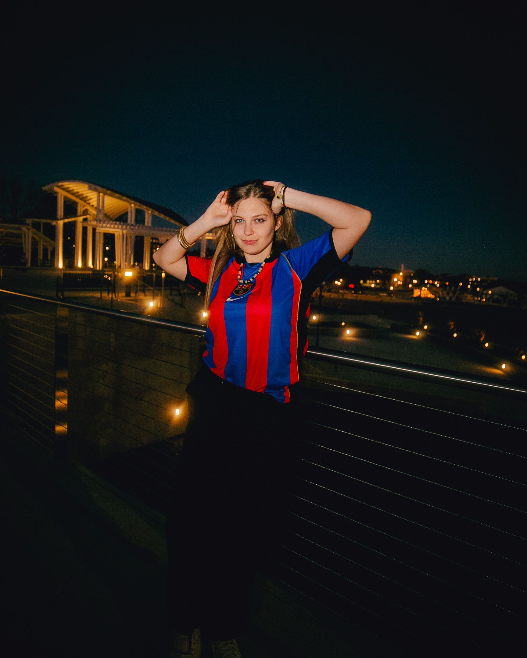 Woman in red and blue striped shirt with hands on head, standing outside at night.