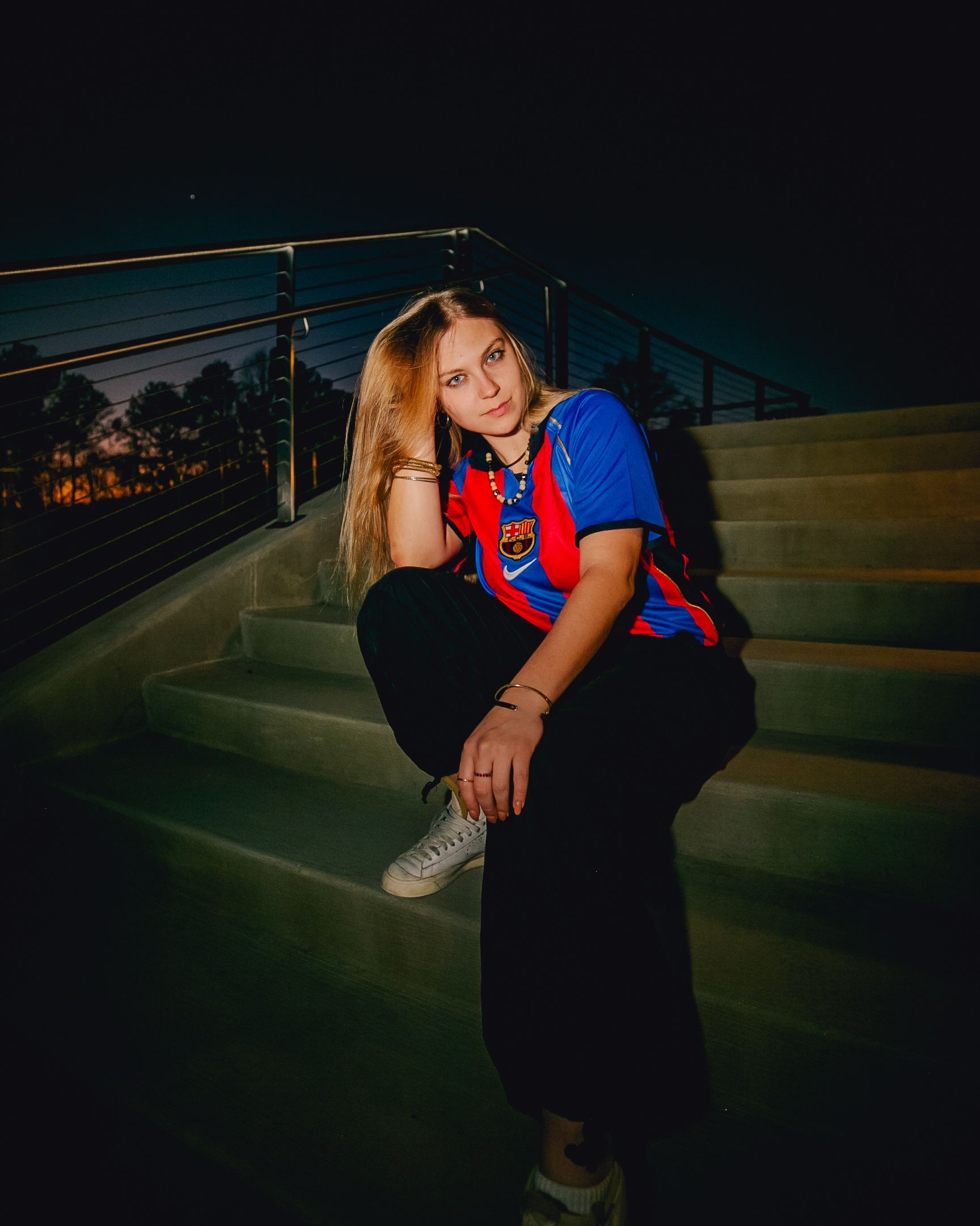 Woman sitting on concrete steps at night wearing a colorful shirt, black pants, and sneakers.