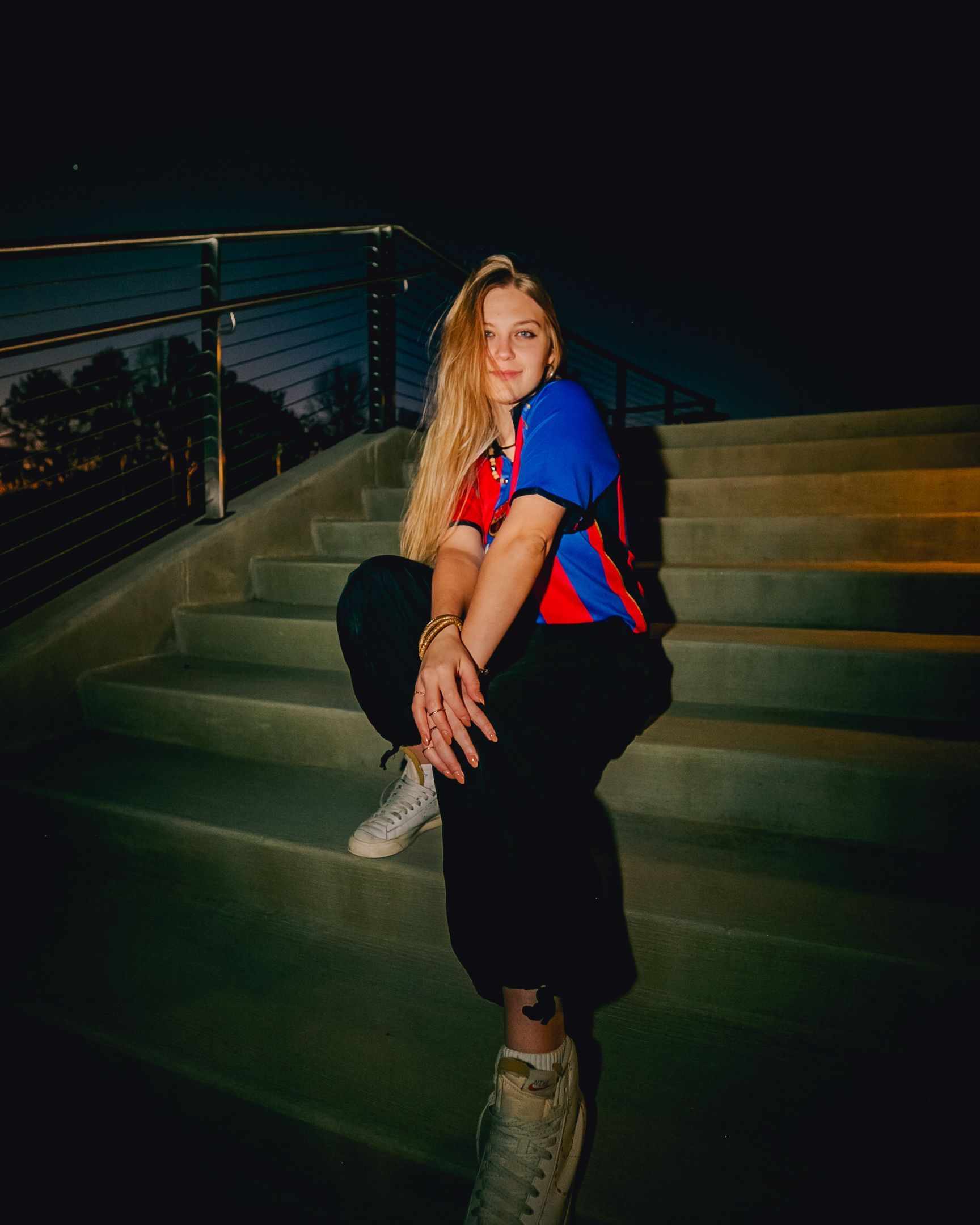 Woman sitting on outdoor steps at night wearing a colorful shirt and black pants.