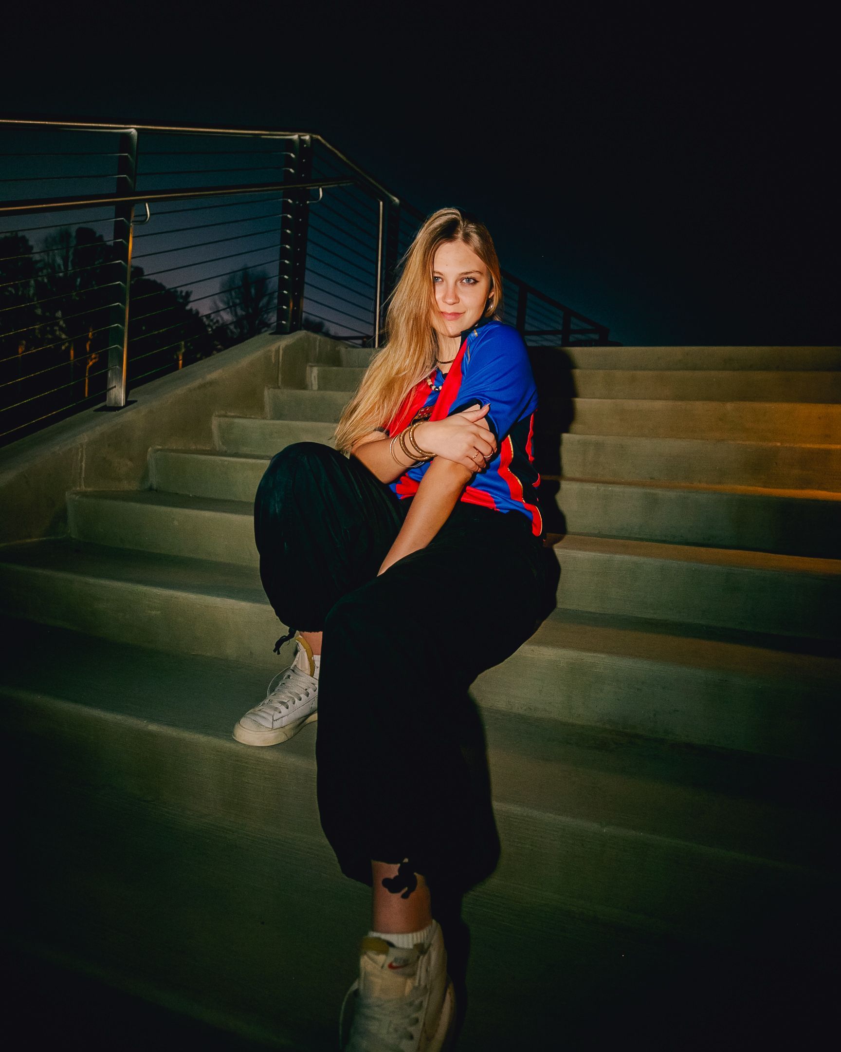 Woman sitting on outdoor steps at night, wearing a blue and red top and black pants.