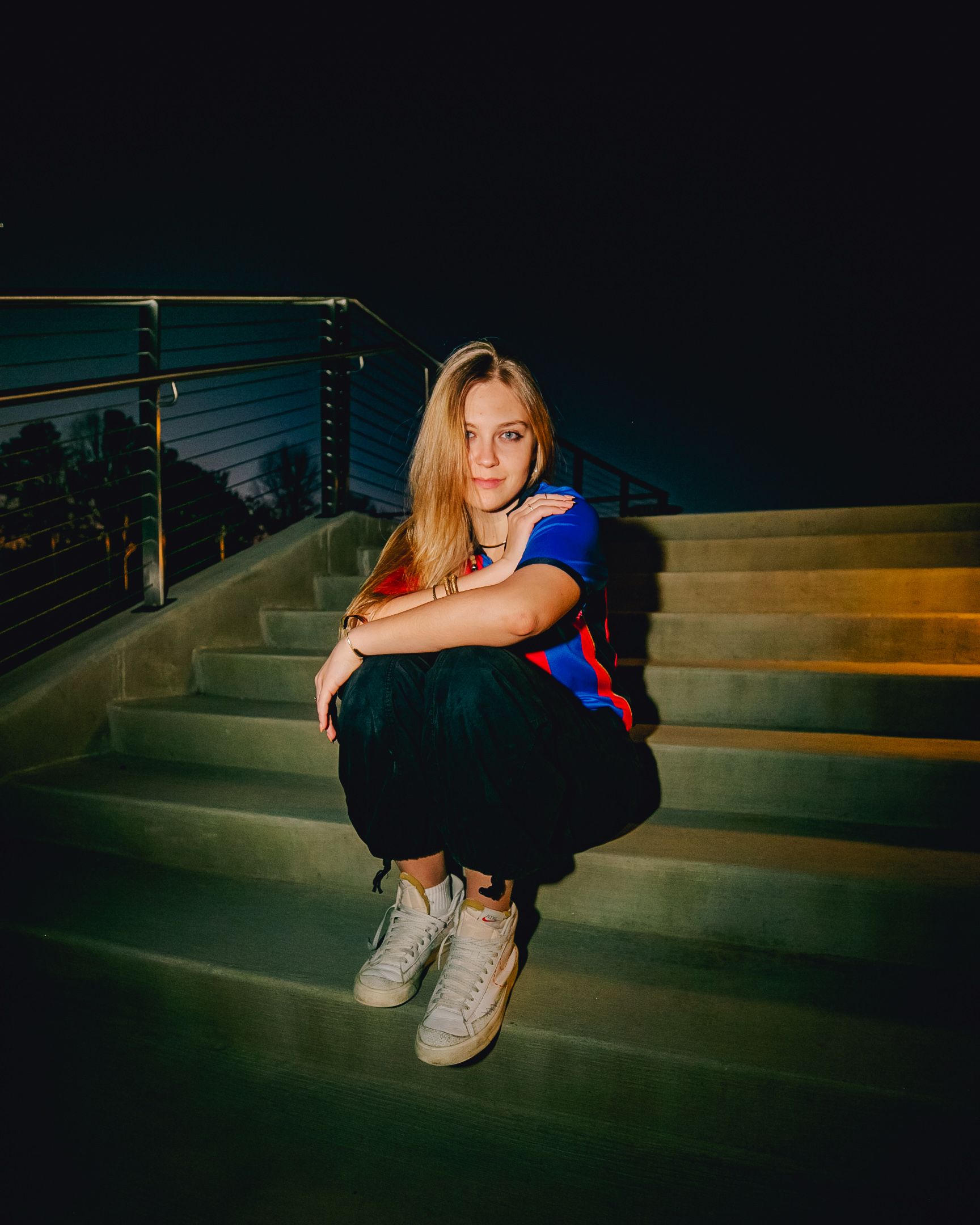 Woman sitting on steps at night, arms crossed. Wearing a blue shirt, black pants, and sneakers.