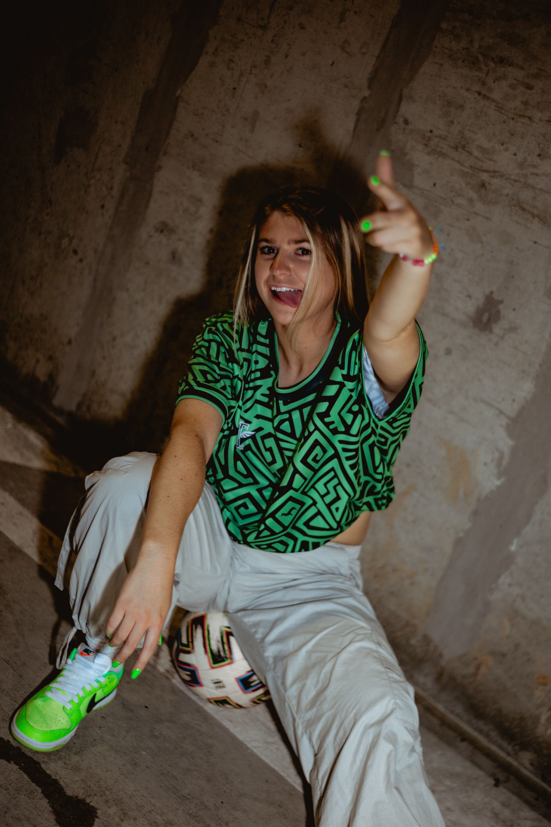 Woman seated with a soccer ball, wearing green patterned shirt, giving a hand gesture, in concrete tunnel.