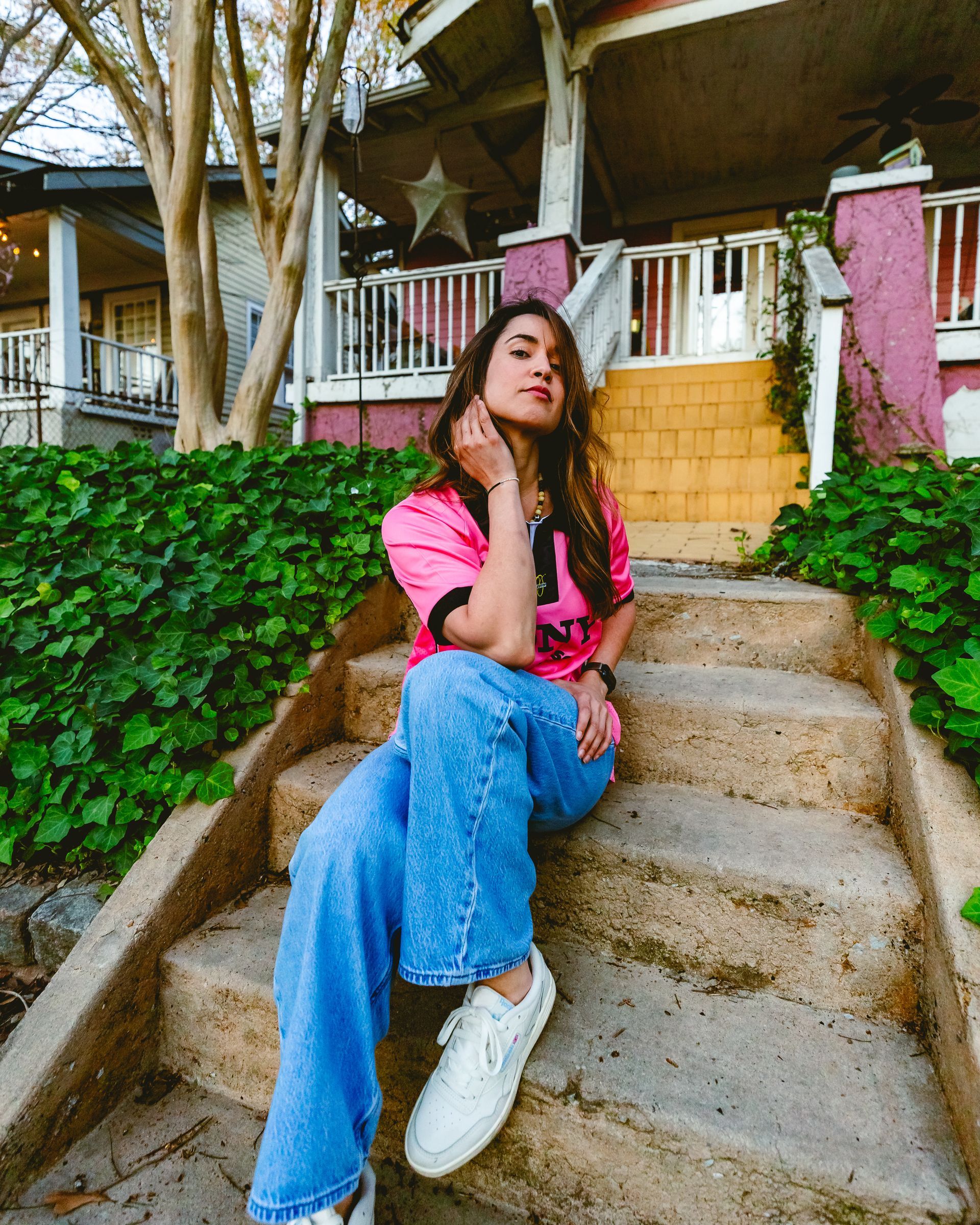 Woman in pink shirt and jeans sits on steps outside a house with green foliage.