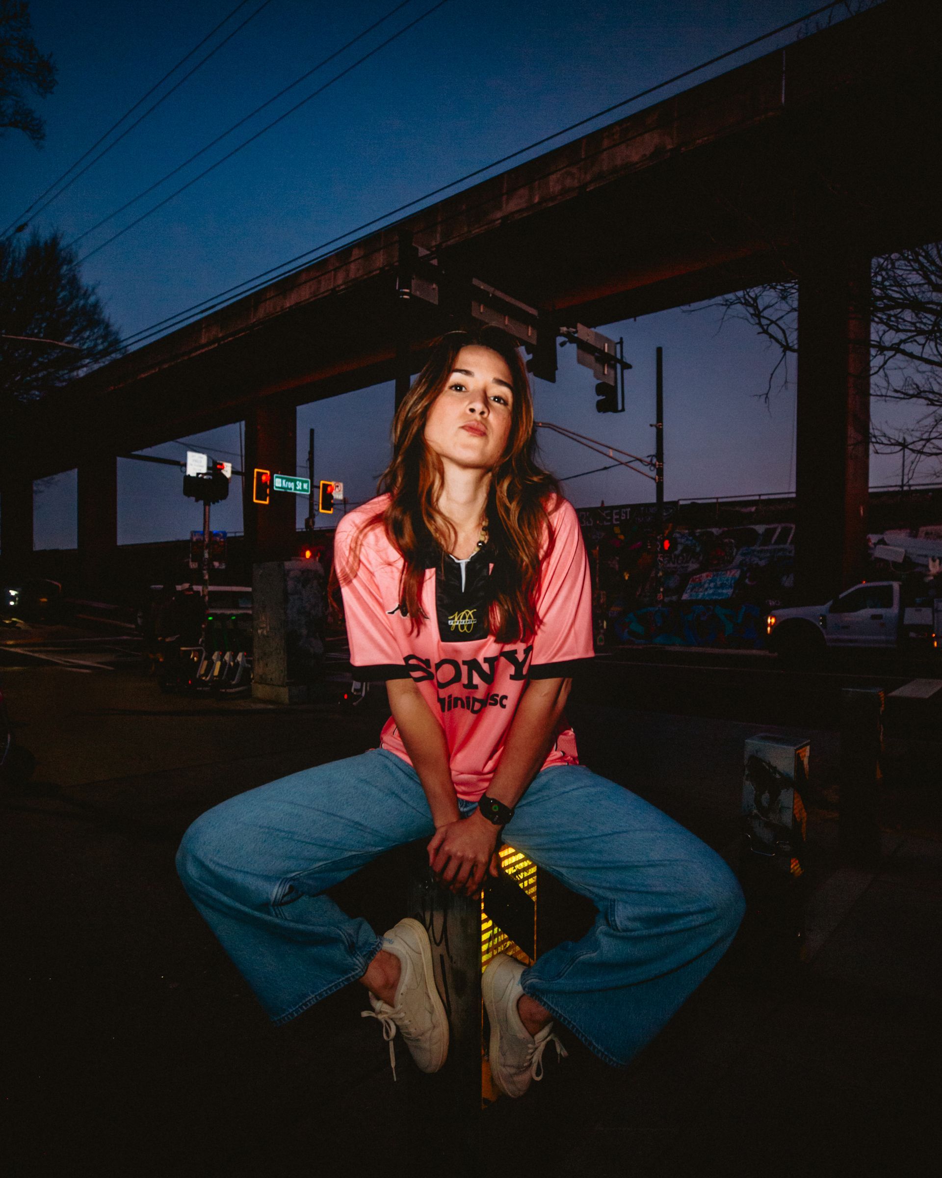 Woman sitting with legs spread, under a bridge at dusk, wearing a pink shirt, blue jeans, and sneakers.