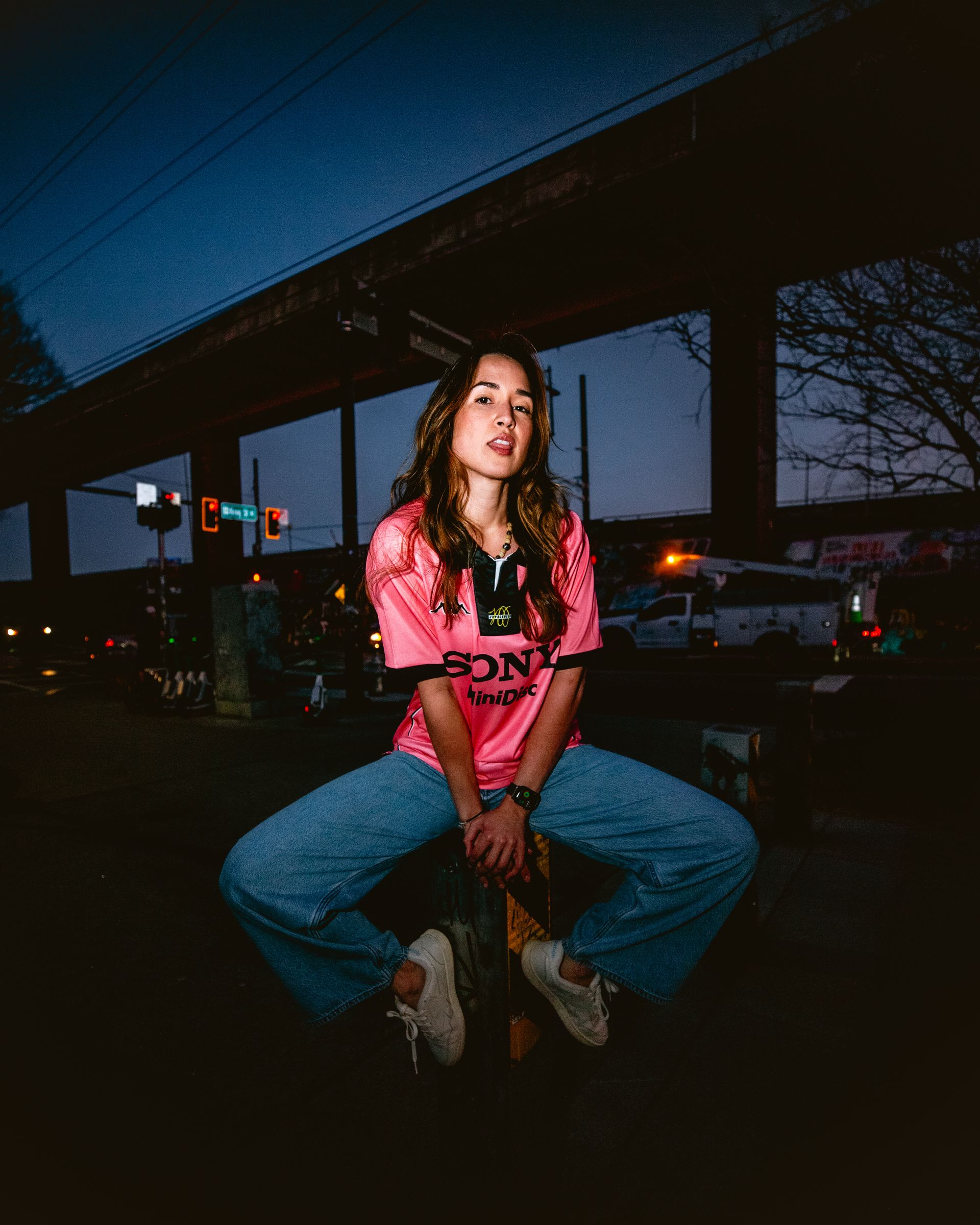 Woman in pink shirt and jeans squats under a bridge at dusk, urban background.