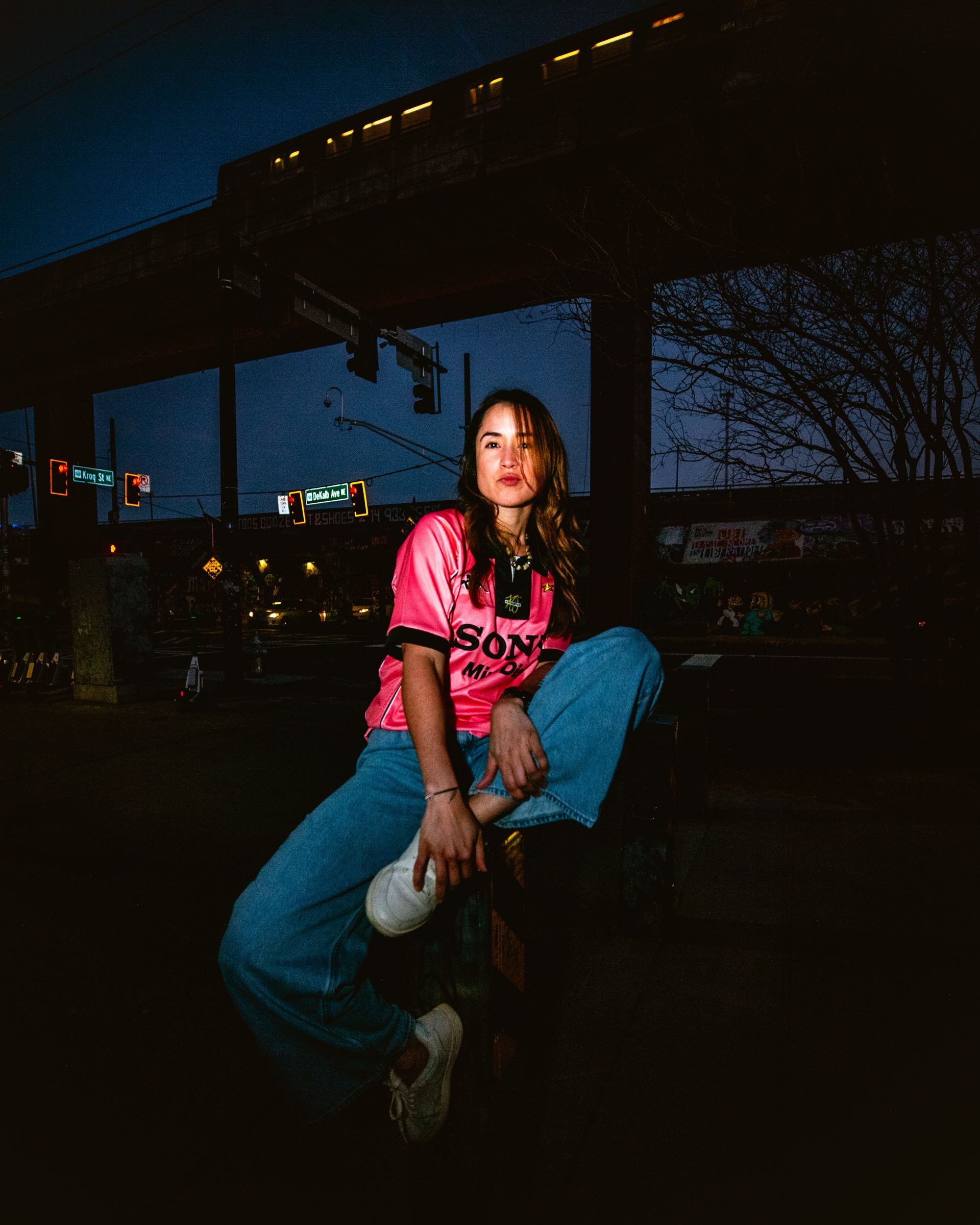 Woman in pink jersey and jeans sitting outdoors at night, holding a water bottle.