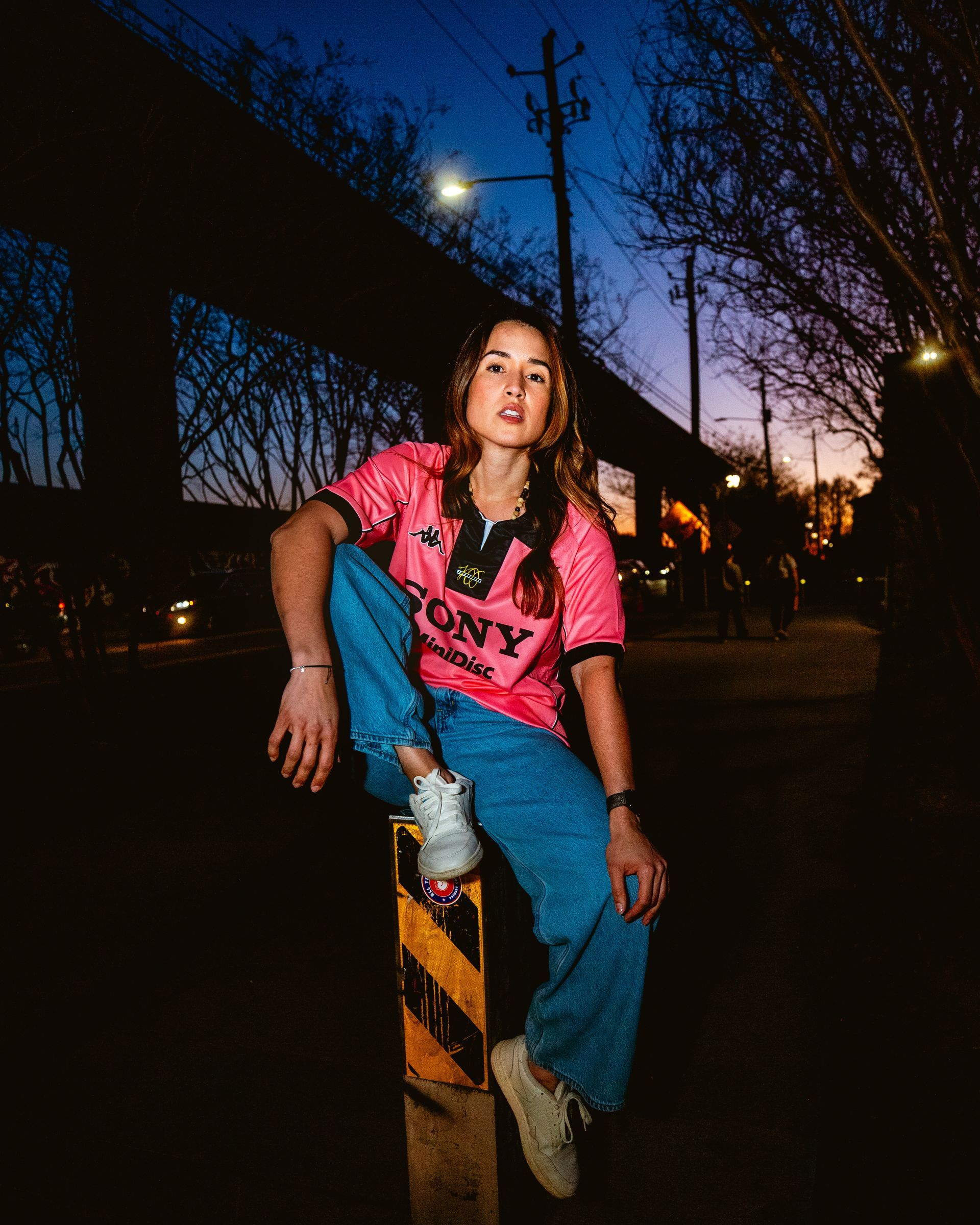 Woman in pink jersey and jeans sits on a street bollard at dusk.
