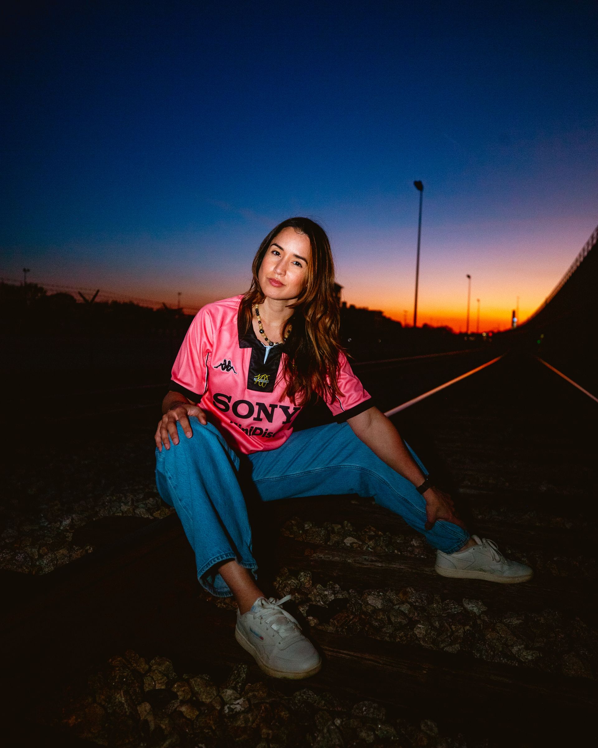 Woman sitting on ground, wearing a pink jersey, blue jeans, and sneakers, with a sunset background.
