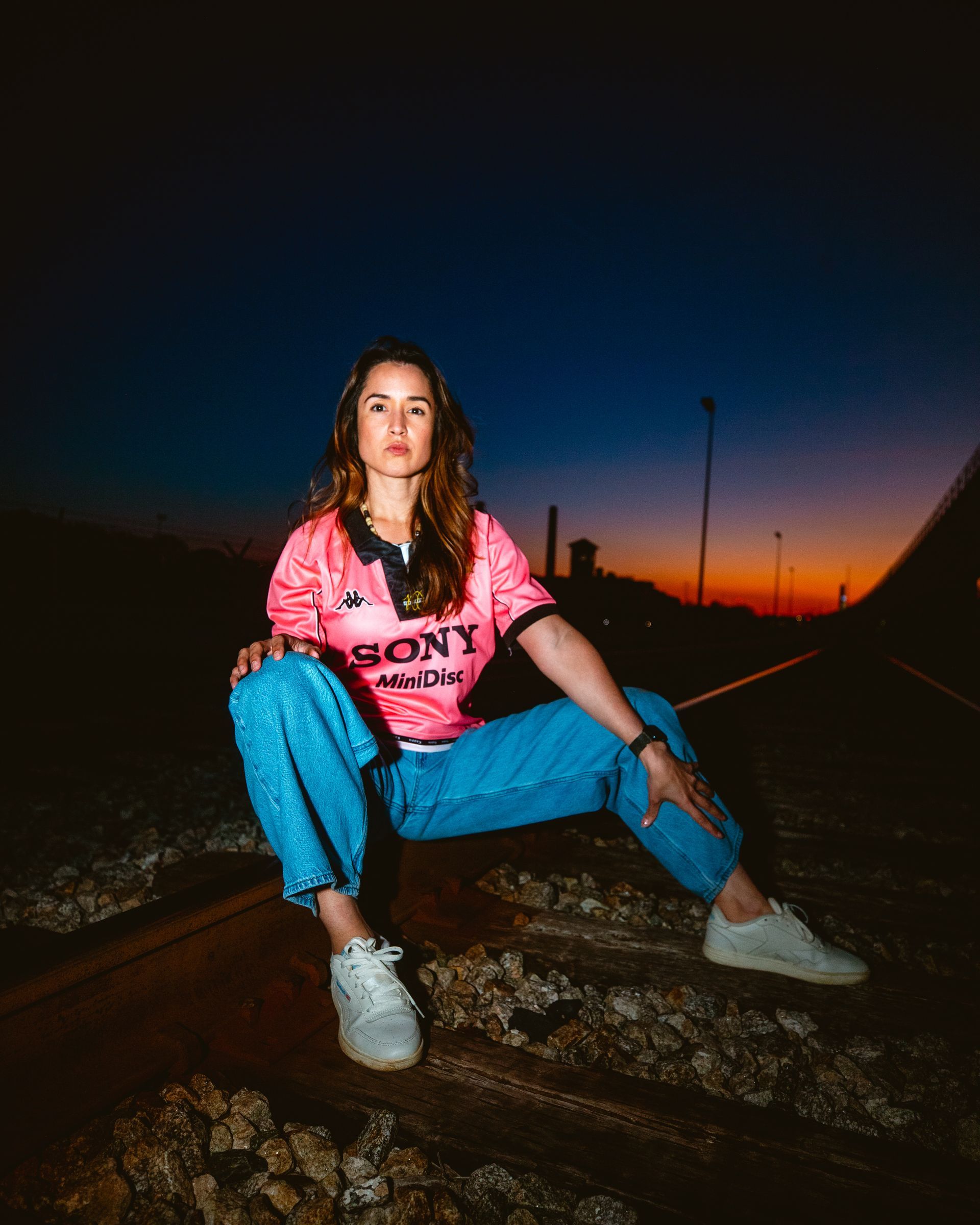 Woman in pink jersey and blue jeans sits on tracks at sunset.