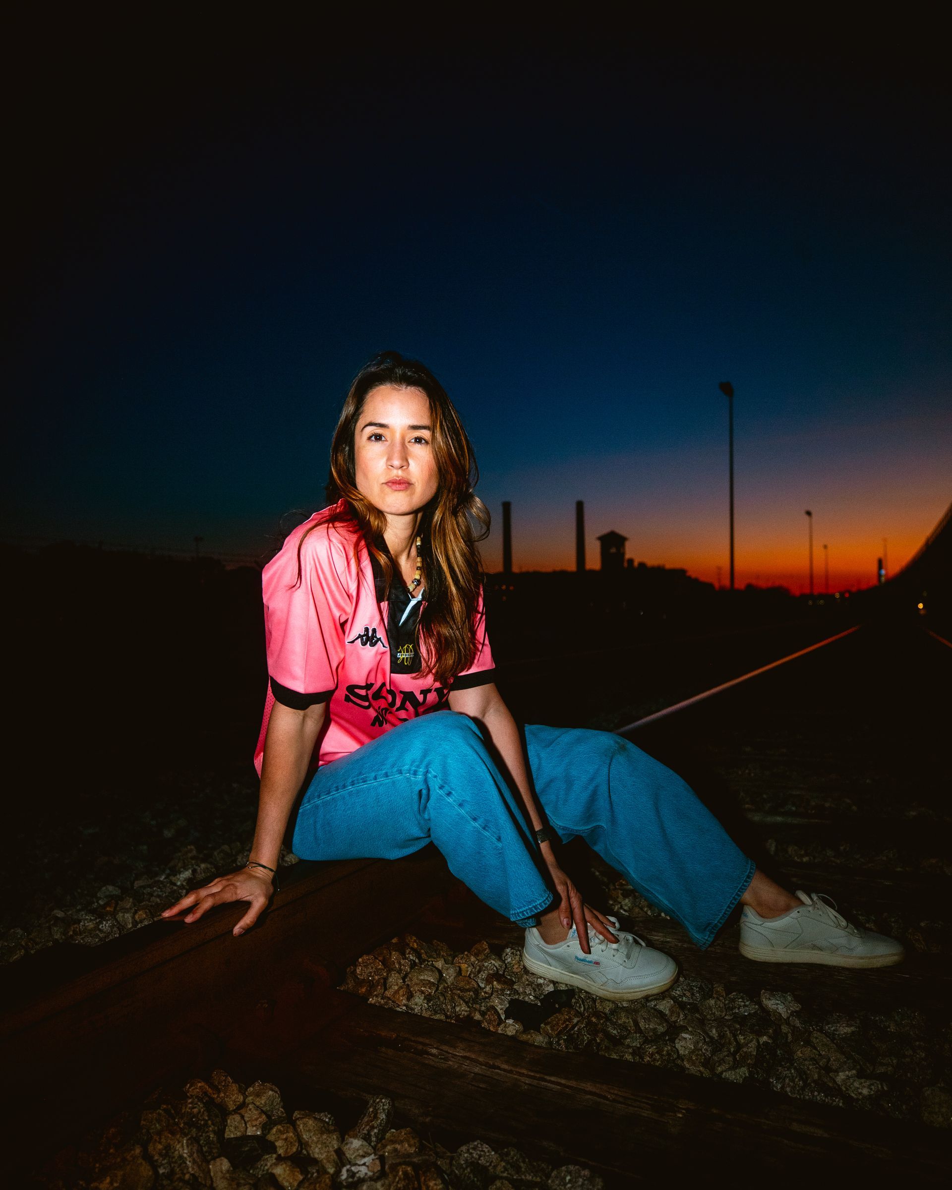 Woman in pink jersey and blue jeans sits on train tracks at dusk.