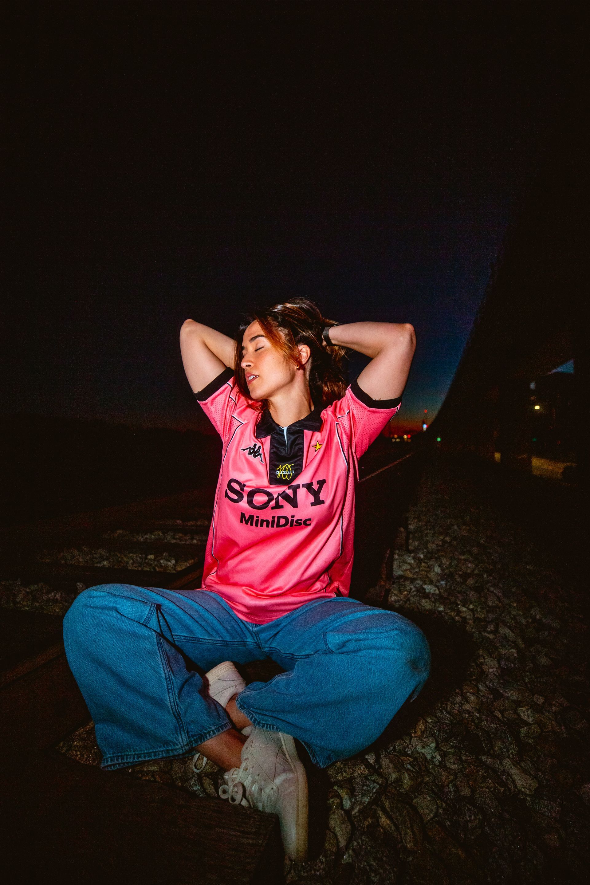 Woman in pink jersey and blue jeans sits cross-legged outdoors at dusk.