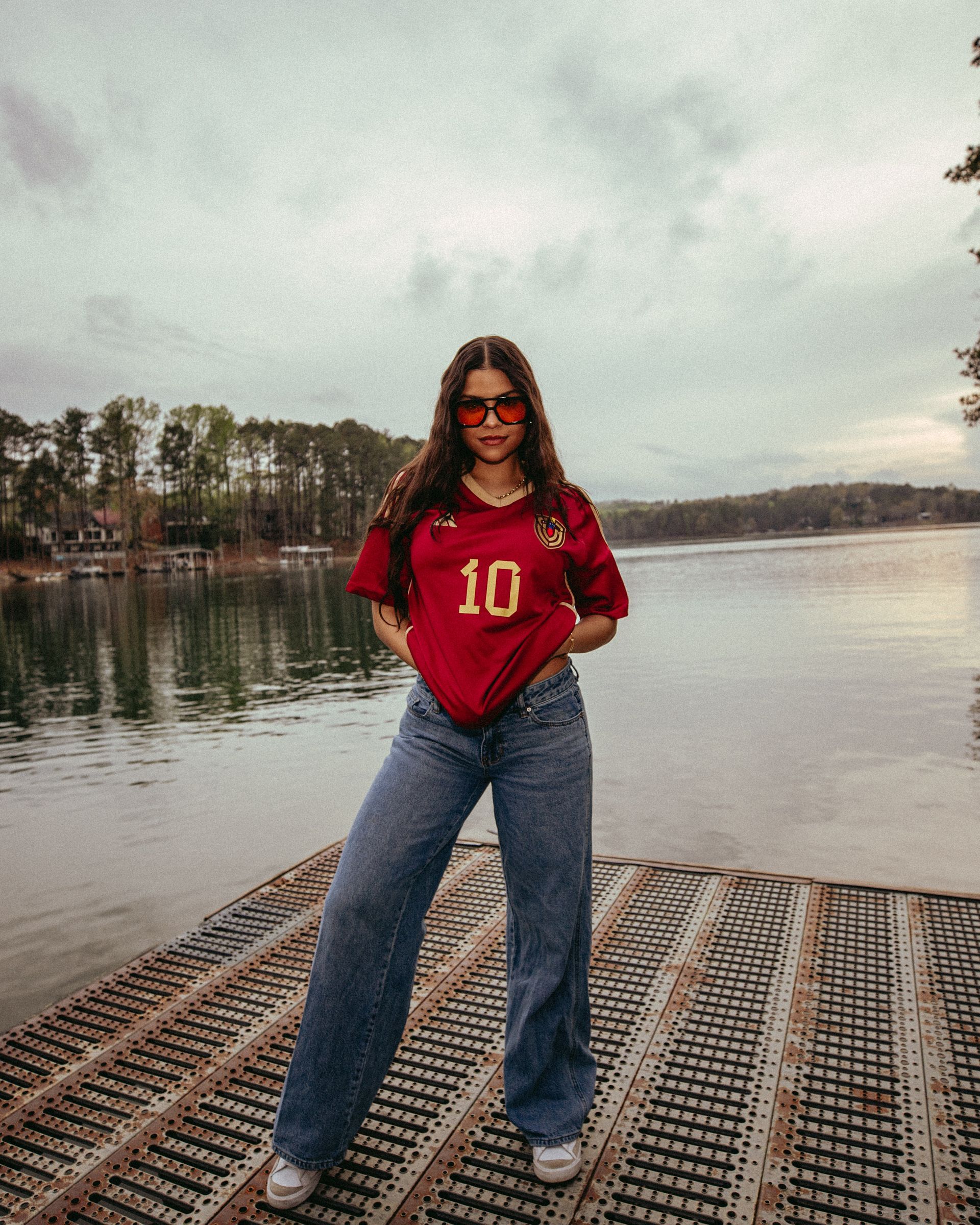 Woman in red jersey and jeans, standing on a dock by a lake, wearing sunglasses, cloudy sky.