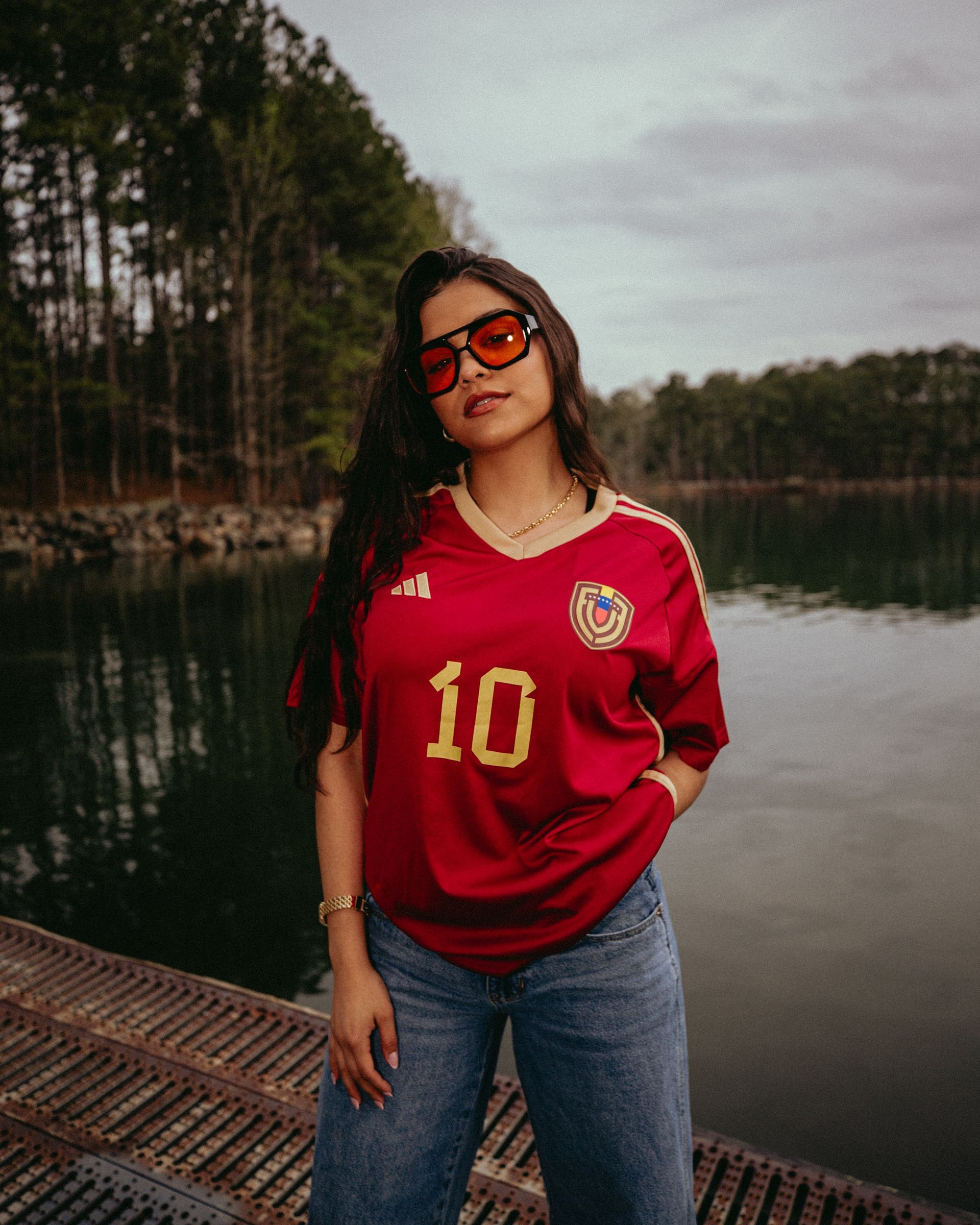 Woman in red jersey and sunglasses stands by water, wearing jeans.