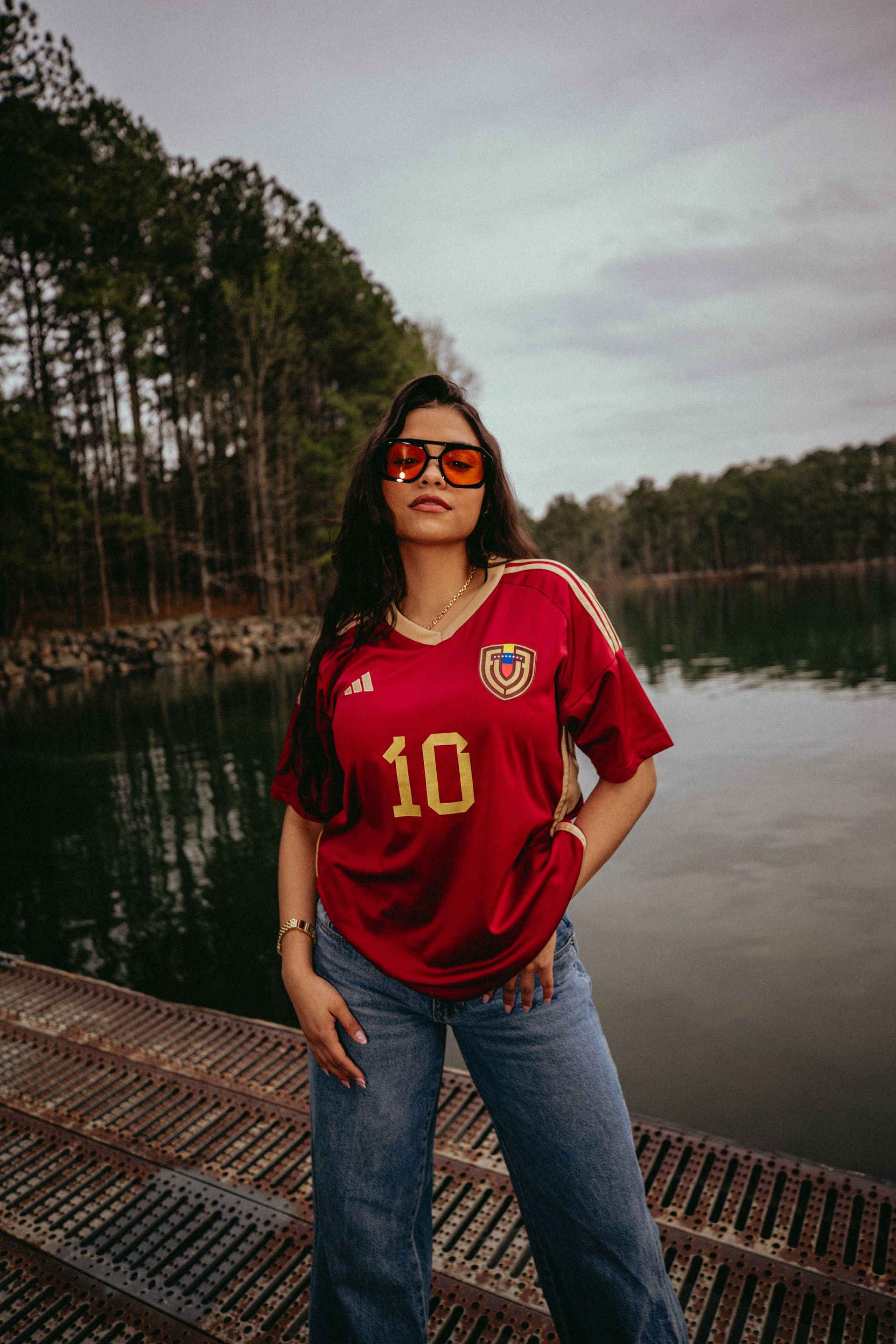 Woman wearing a red jersey with the number 10, sunglasses, and jeans, standing near water.