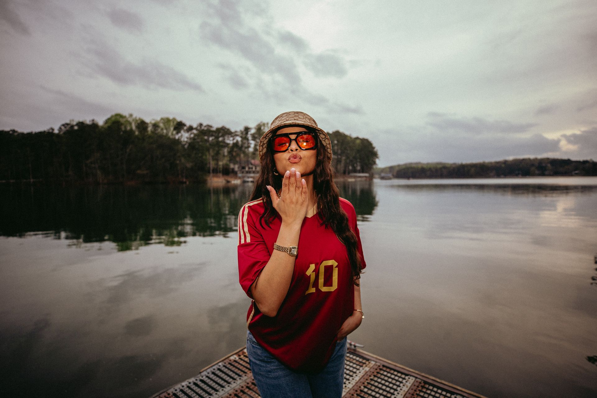 Woman in red jersey blowing a kiss toward the camera, standing by a lake. Overcast sky.