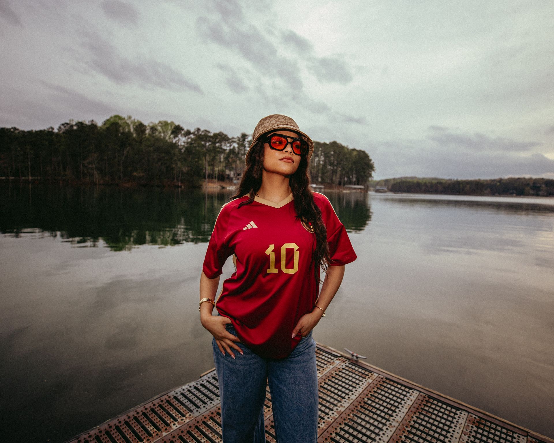 Woman in red jersey and sunglasses poses on a dock overlooking a lake.