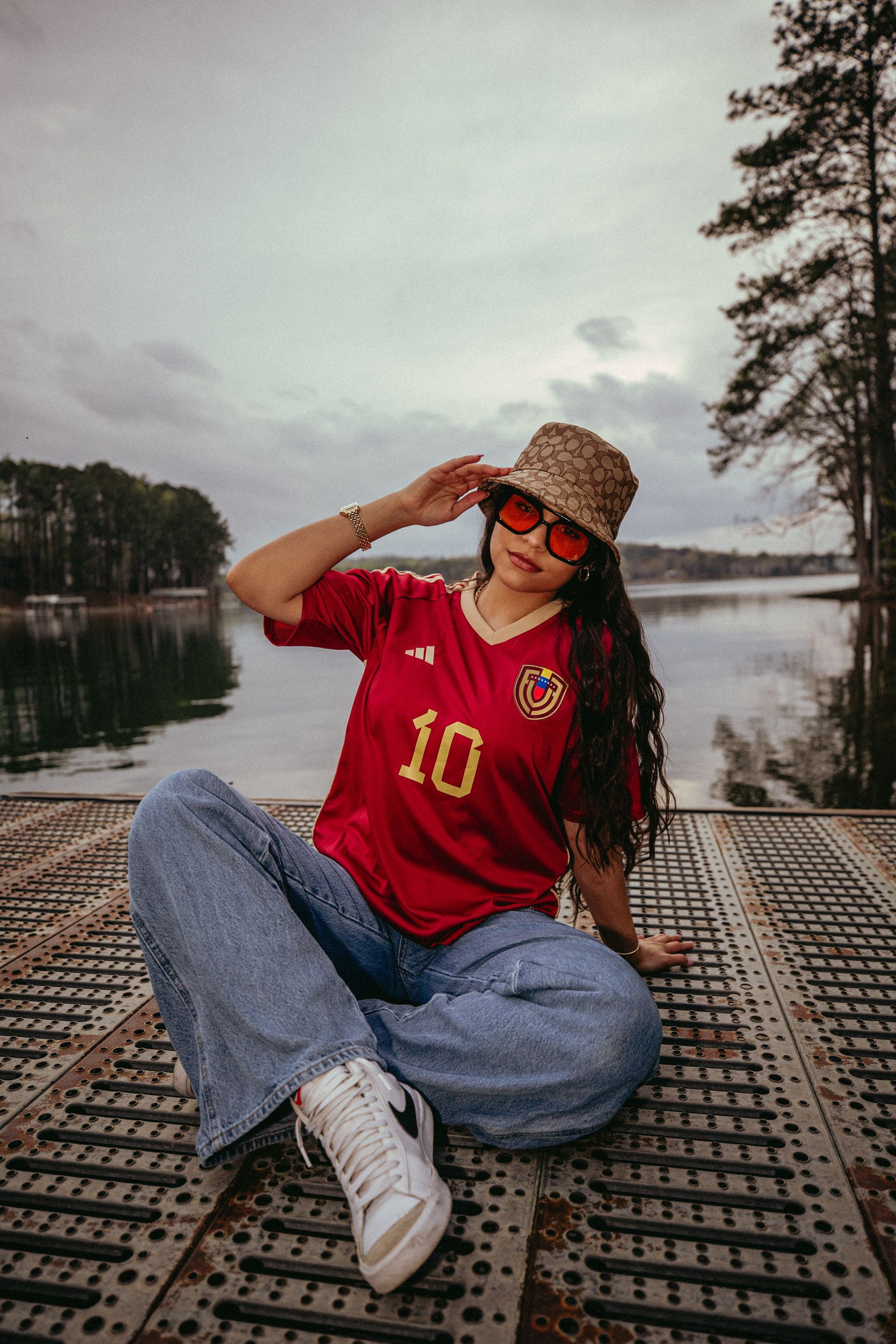 Woman wearing red jersey and hat, sitting by lake.