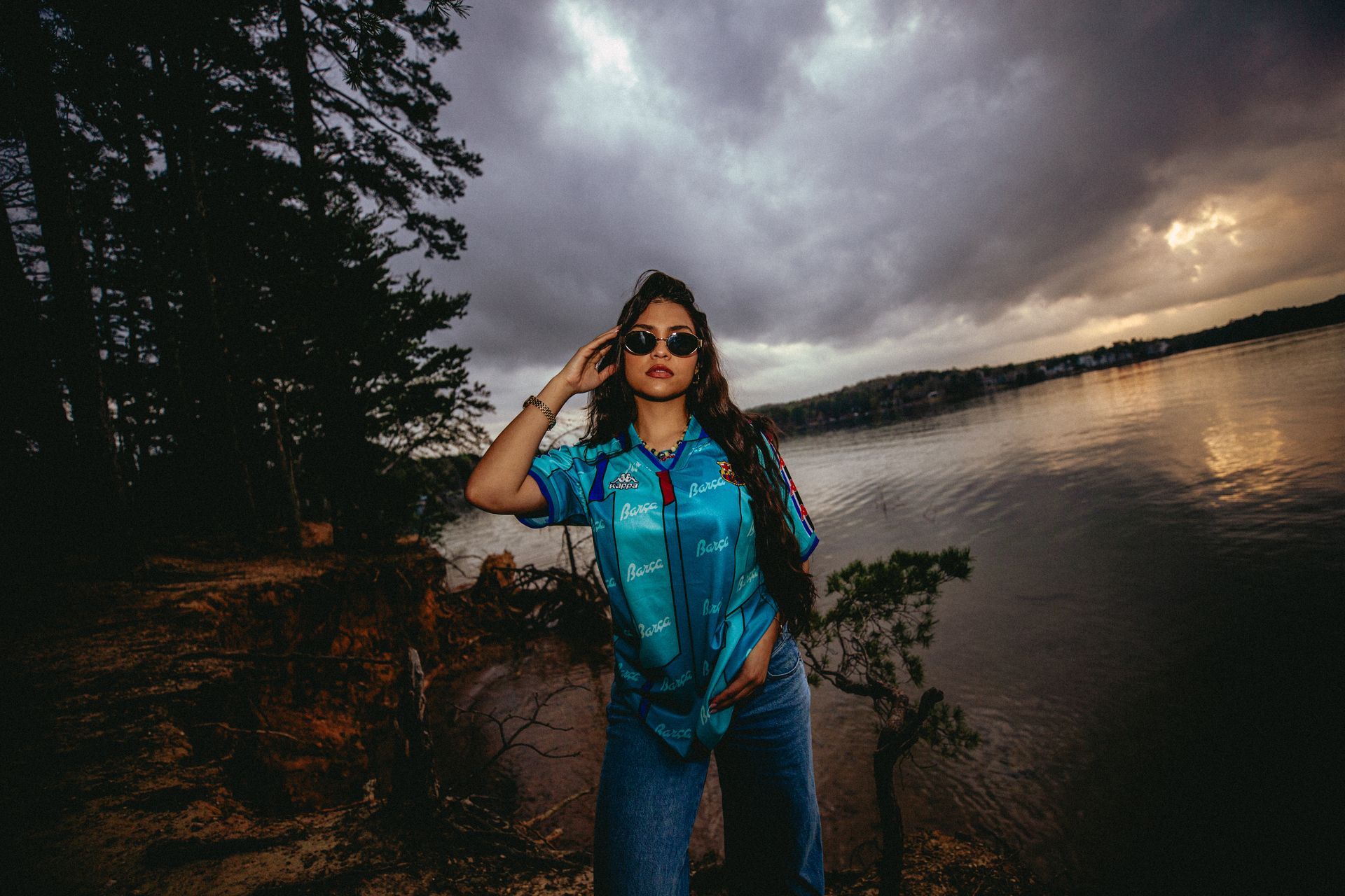 Woman in blue shirt and jeans by a lake, wearing sunglasses, under a cloudy sky.