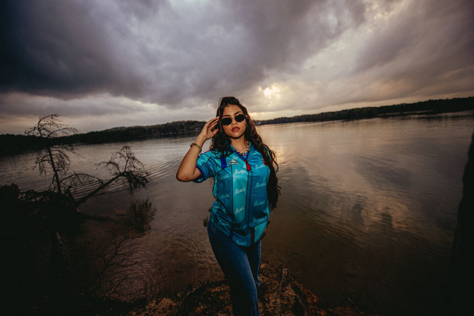 Woman in sunglasses and blue shirt stands by water, stormy sky in background.