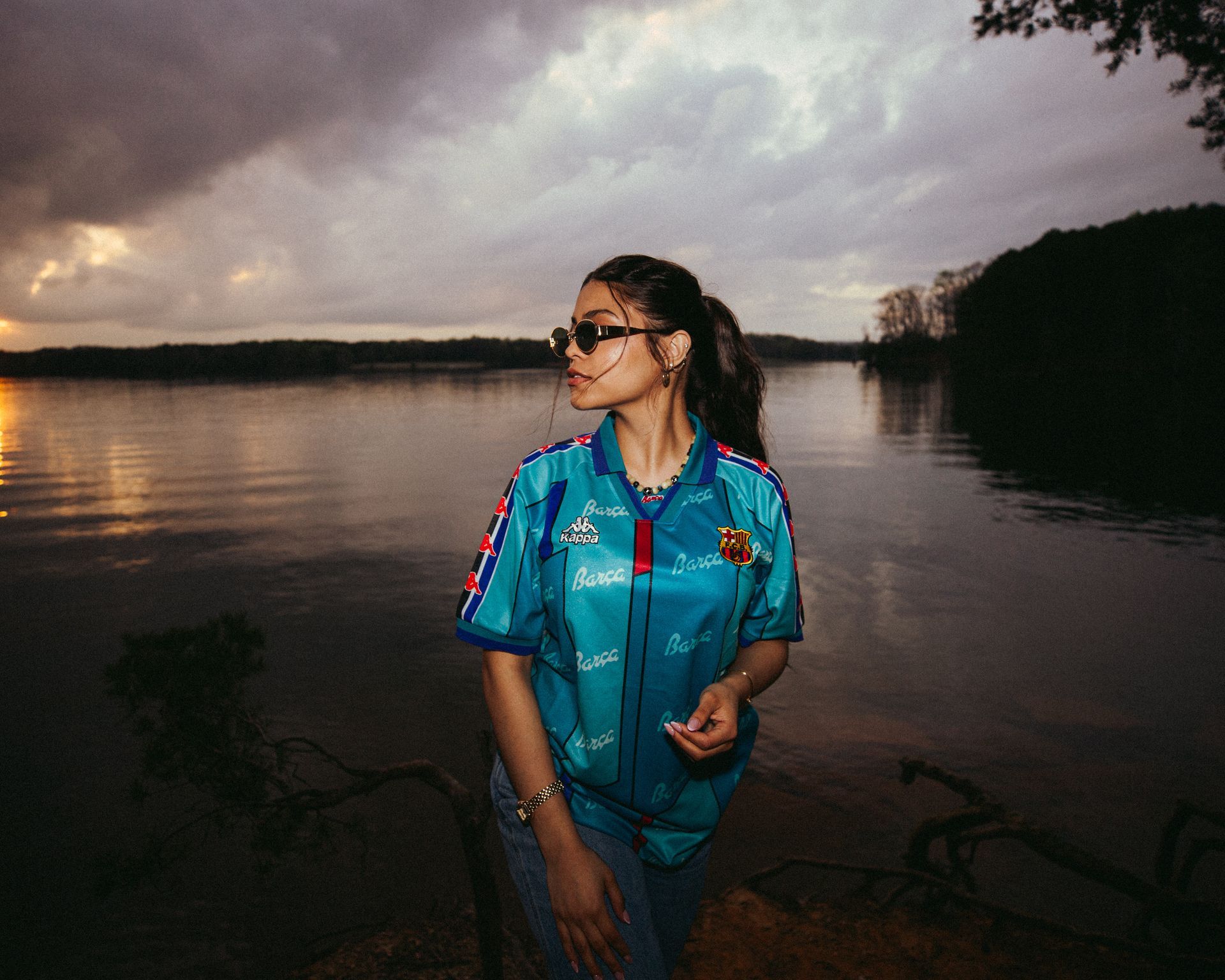 Person wearing sunglasses and blue shirt stands by a lake under a cloudy sky.