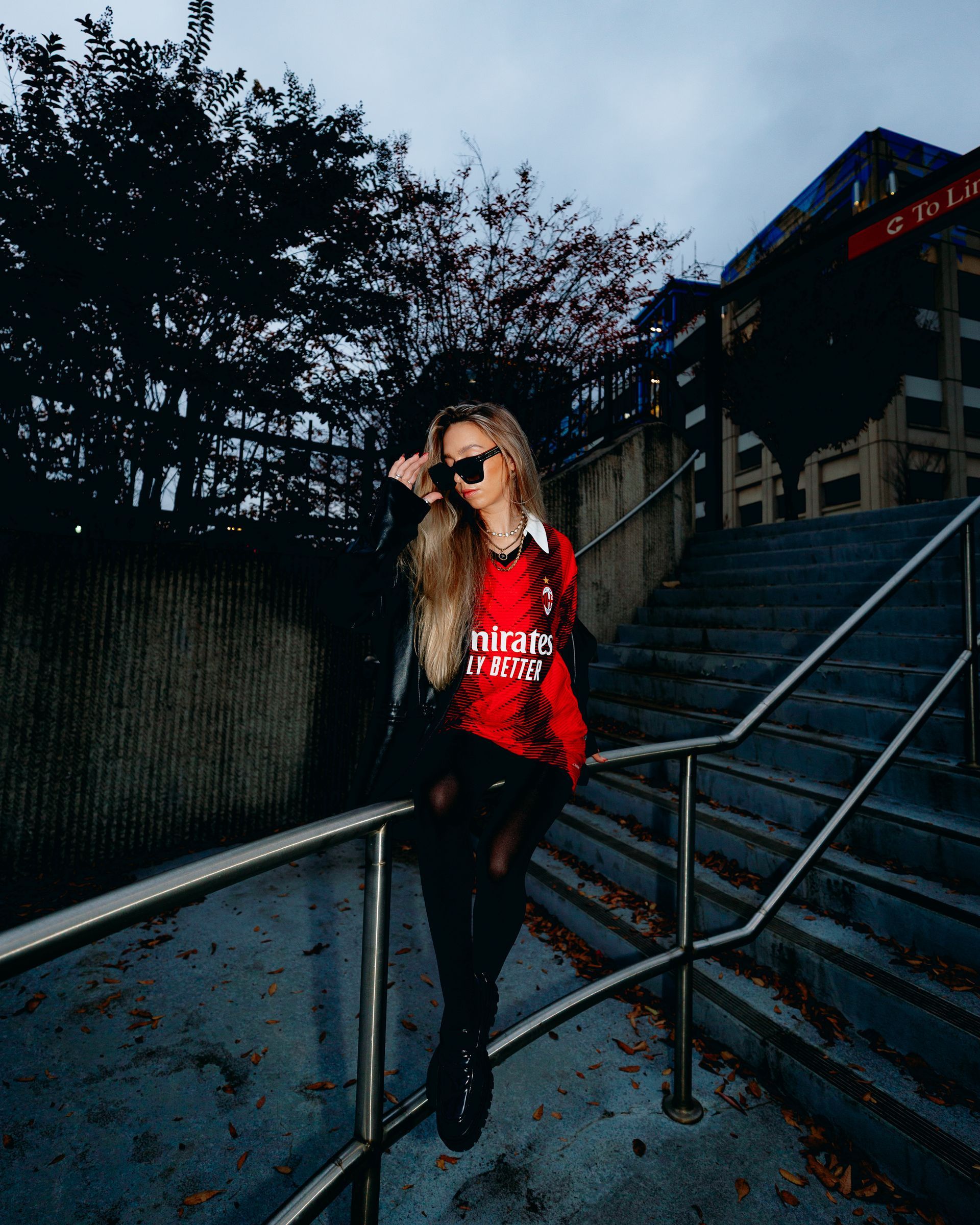 Woman in sunglasses, red shirt, and black boots posing on outdoor stairs with a building backdrop.
