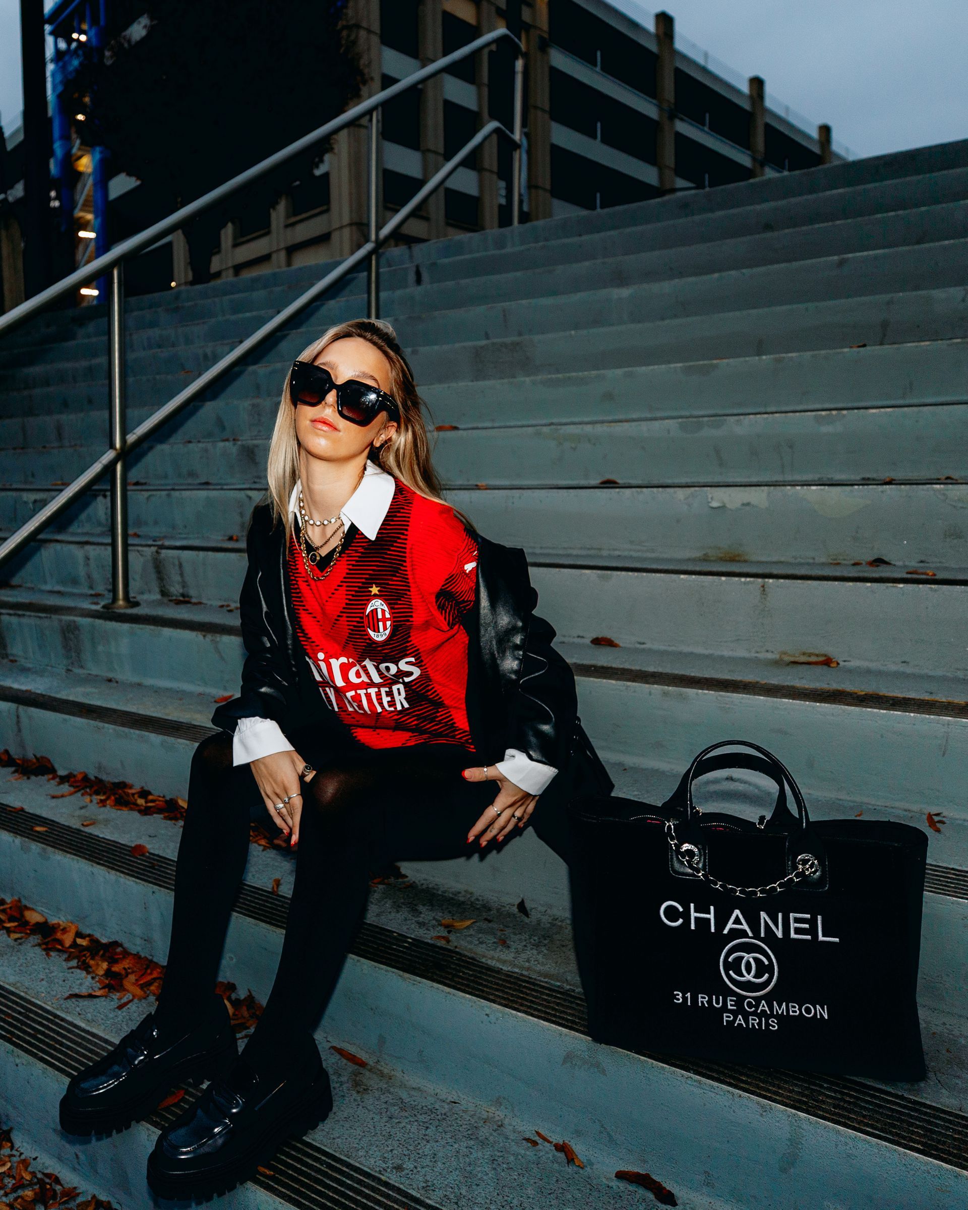 Woman in black outfit and sunglasses, sitting on concrete stairs, with a Chanel bag.