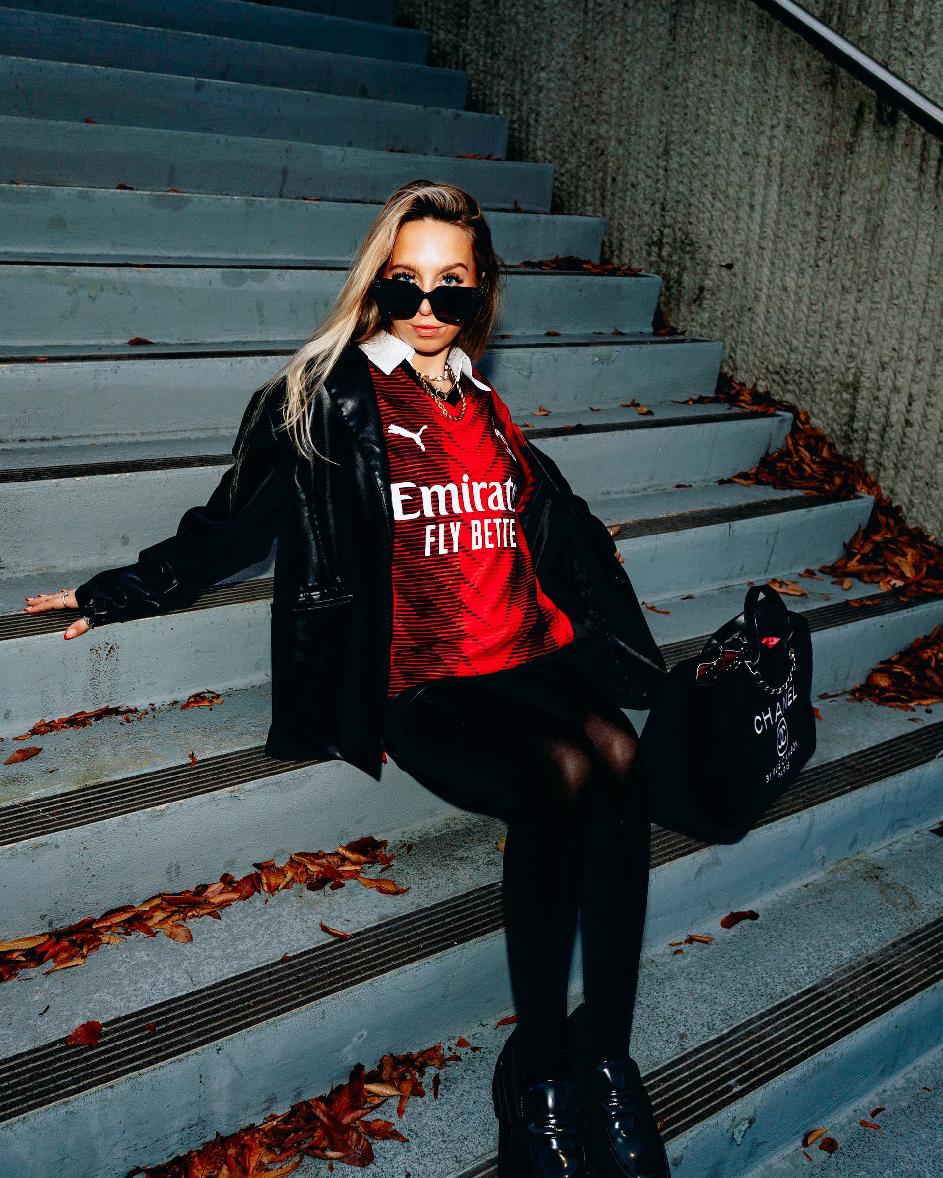 Woman in AC Milan jersey, black jacket, sunglasses, and boots sits on outdoor stairs.
