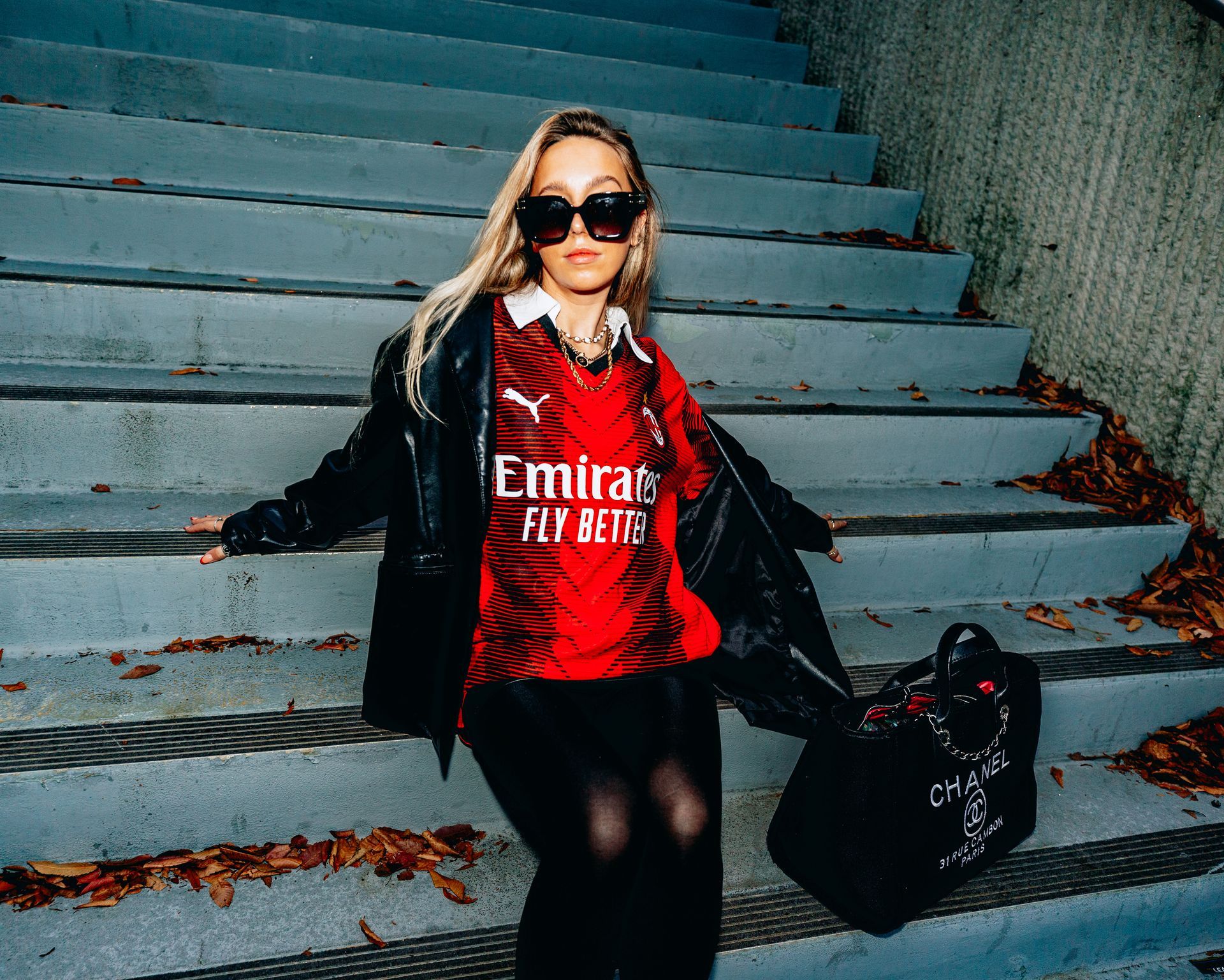 Woman wearing soccer jersey and sunglasses sits on concrete stairs with a black bag.