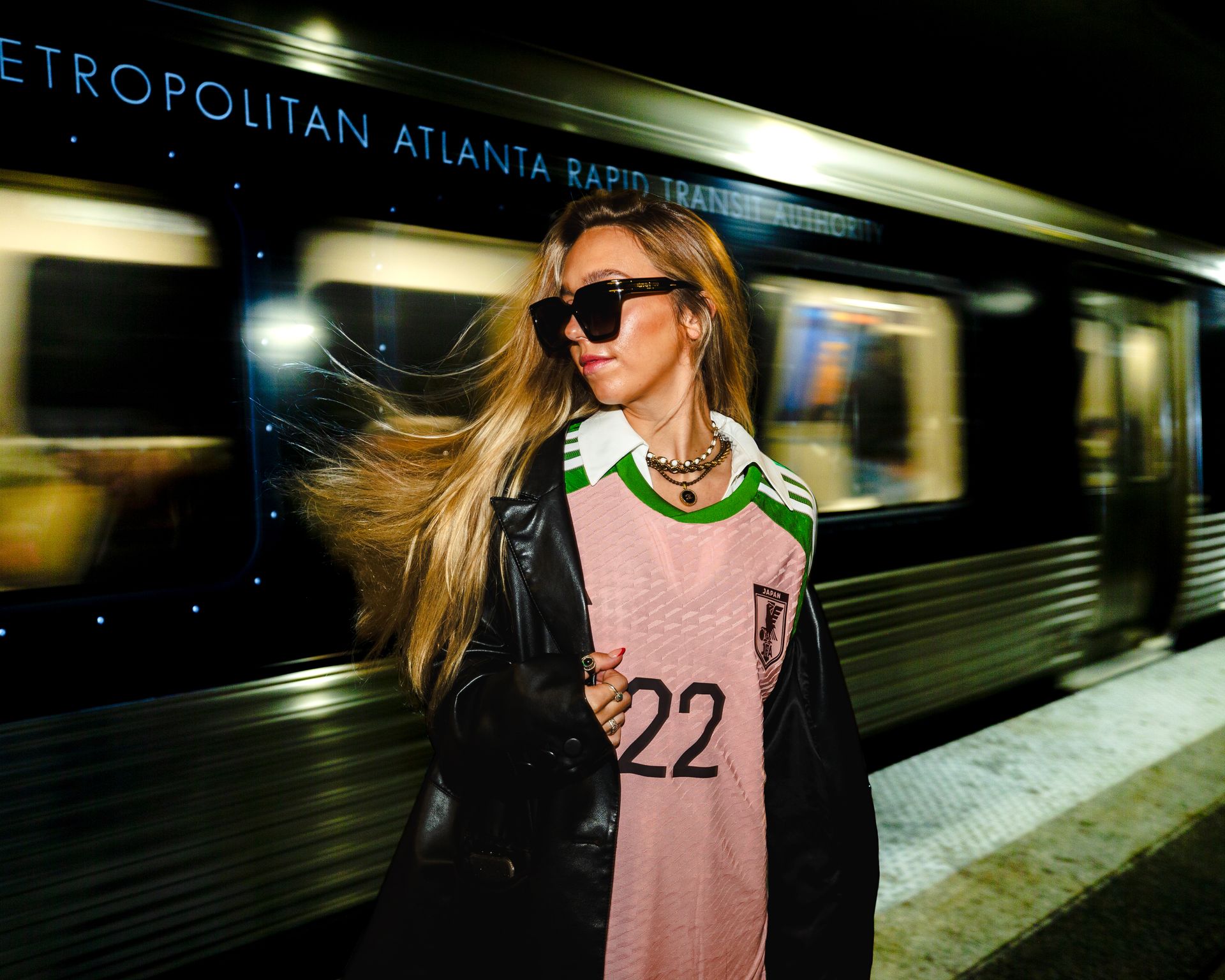 Woman in sunglasses, standing on a subway platform. Train in motion behind her.