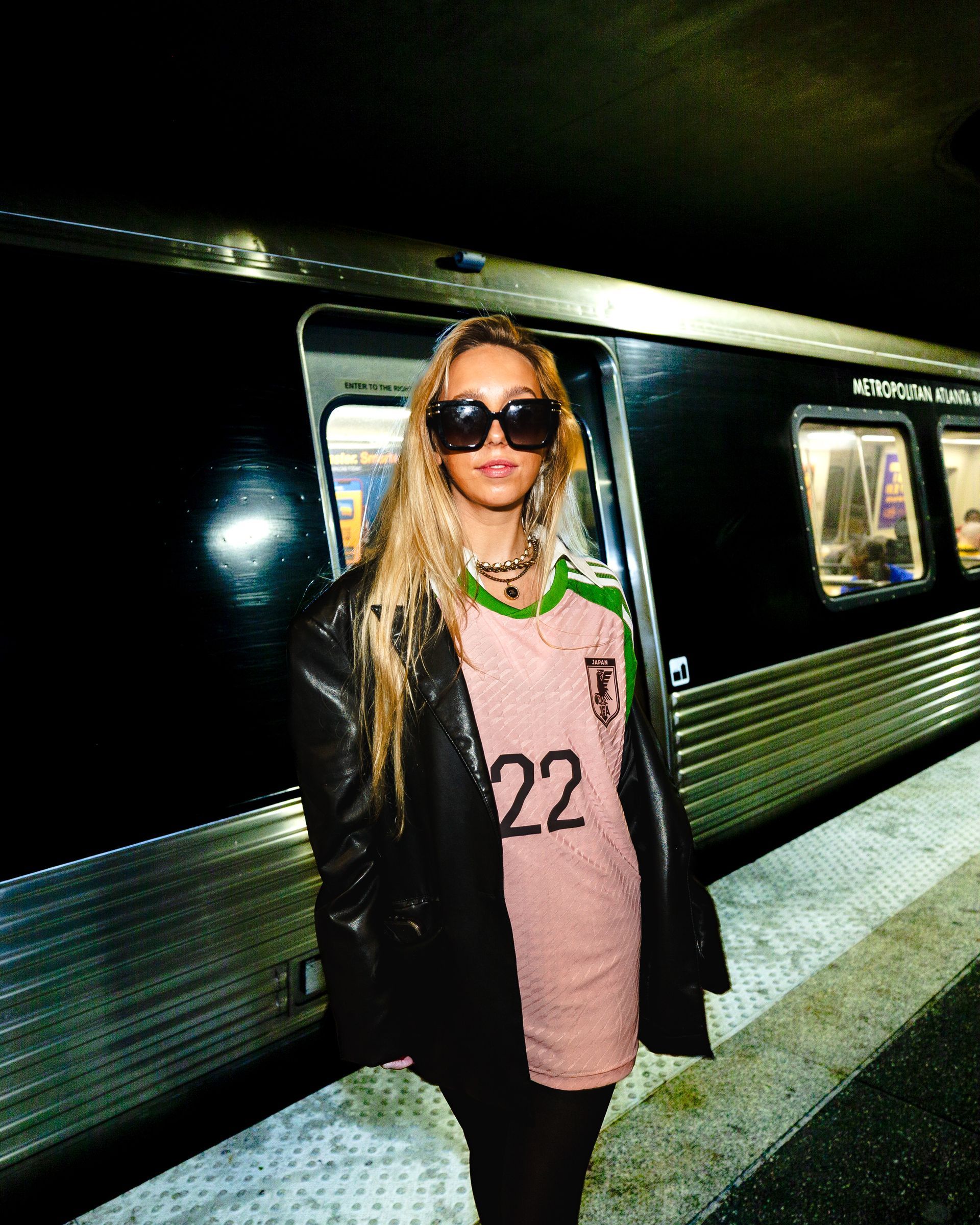 Woman in sunglasses and black jacket stands on a subway platform. She wears a pink jersey with the number 22. Subway car in background.