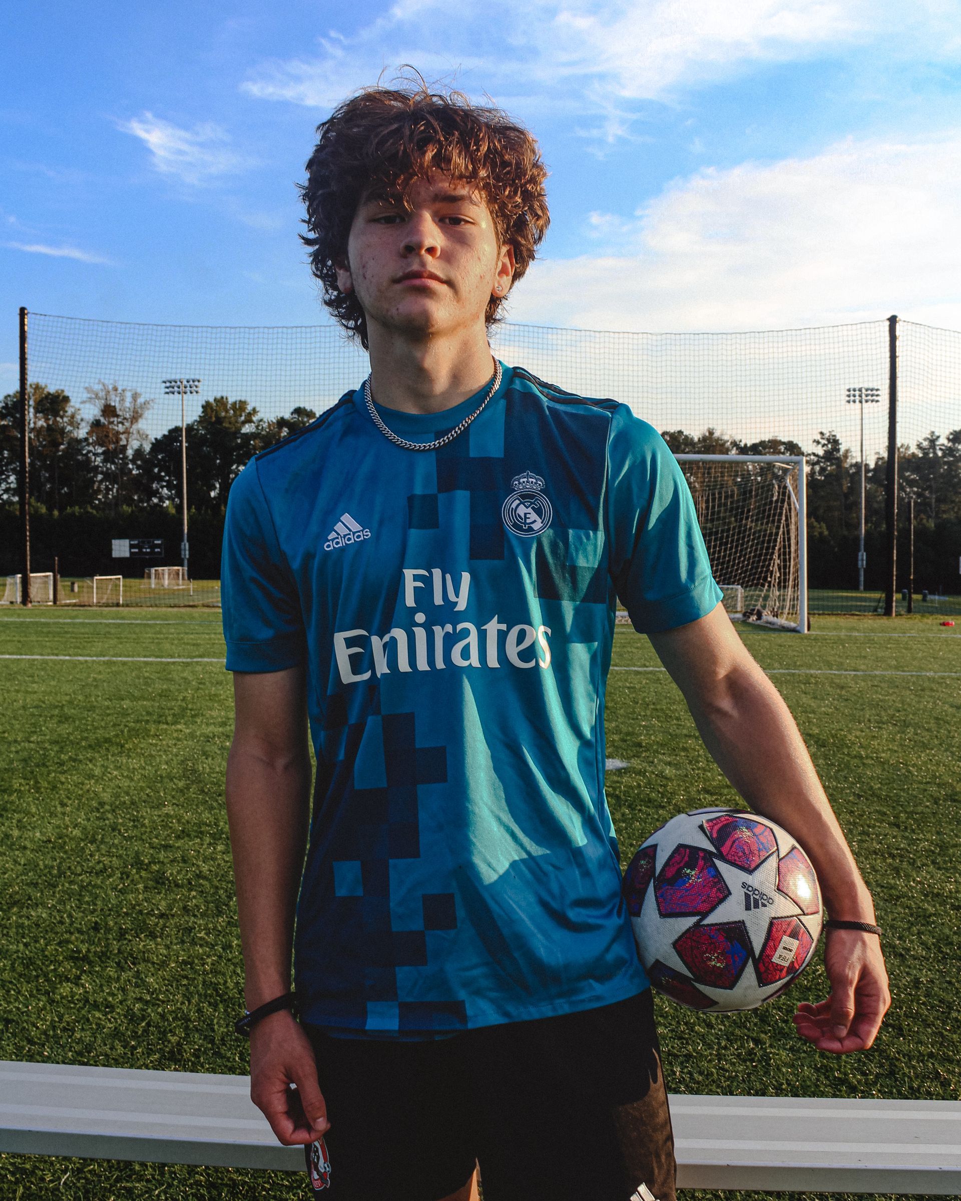 Young person wearing a blue Real Madrid jersey, holding a soccer ball on a field.