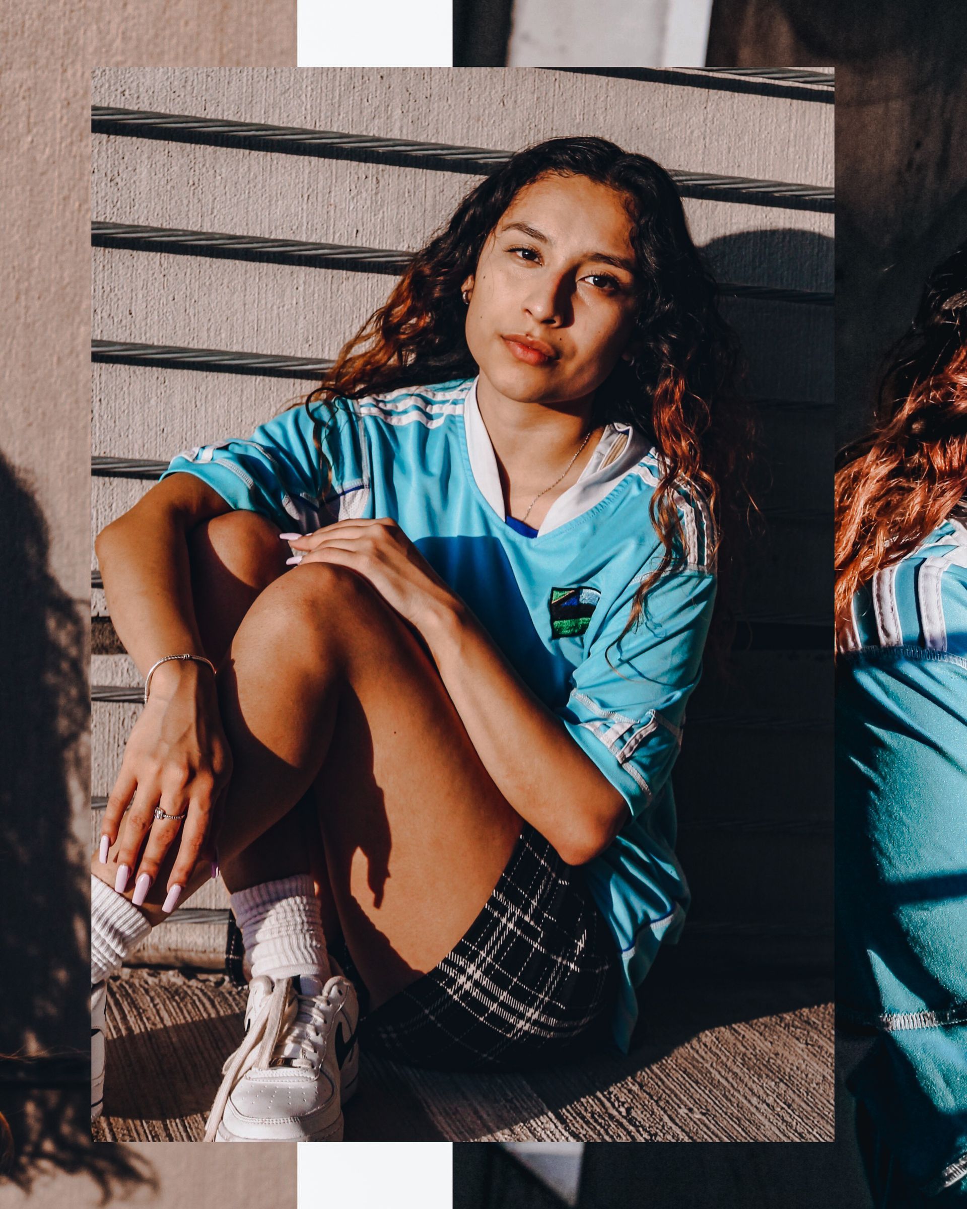 Woman in a light blue jersey and plaid shorts, sitting on stairs, sunny outdoor setting.