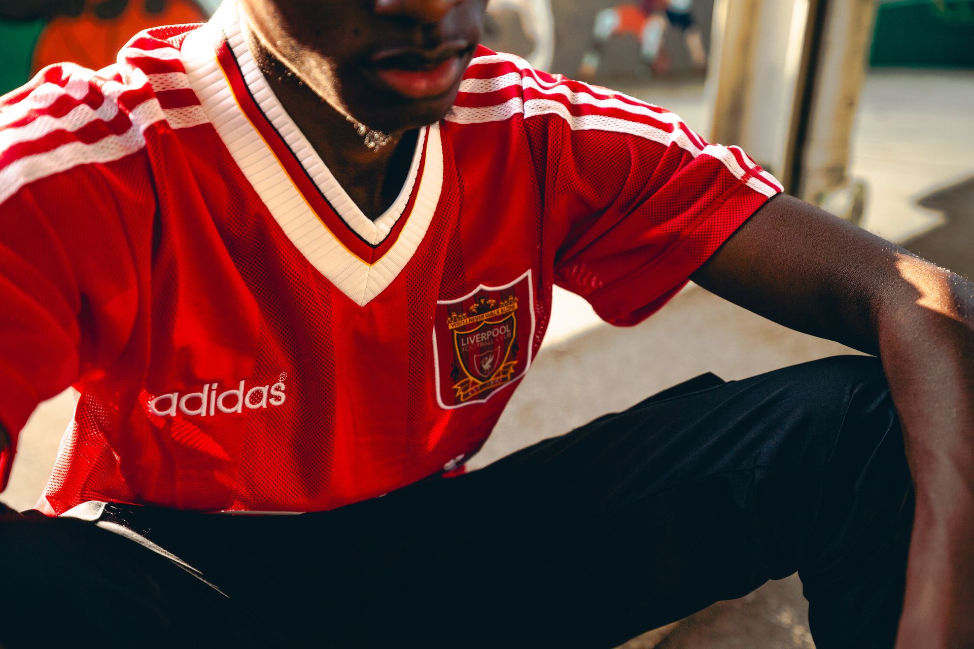 Man wearing a red Liverpool soccer jersey, Adidas logo visible. Sitting, resting in sunlit outdoor area.
