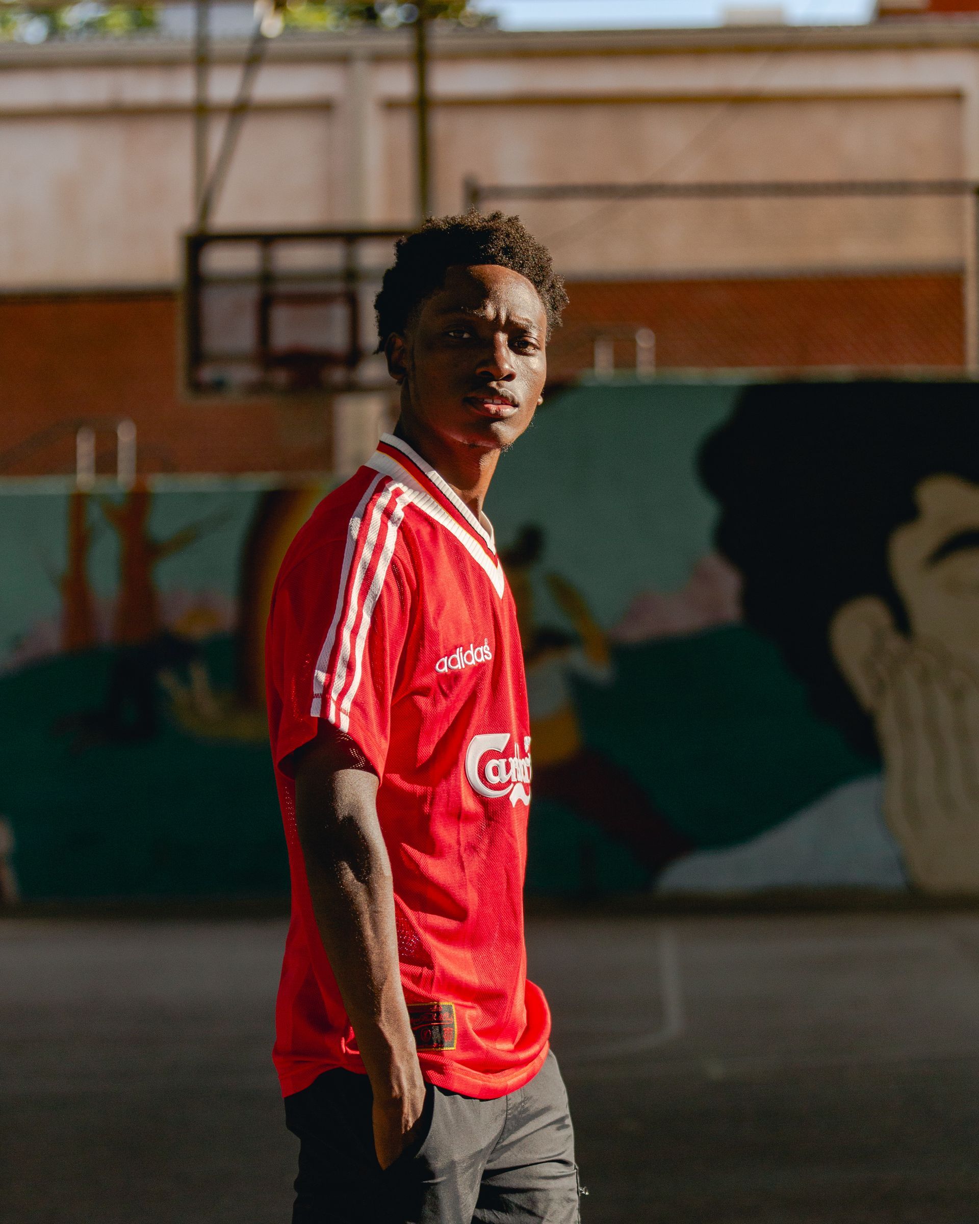 Man in red jersey stands on a basketball court, looking at the camera. A mural and hoop are in the background.