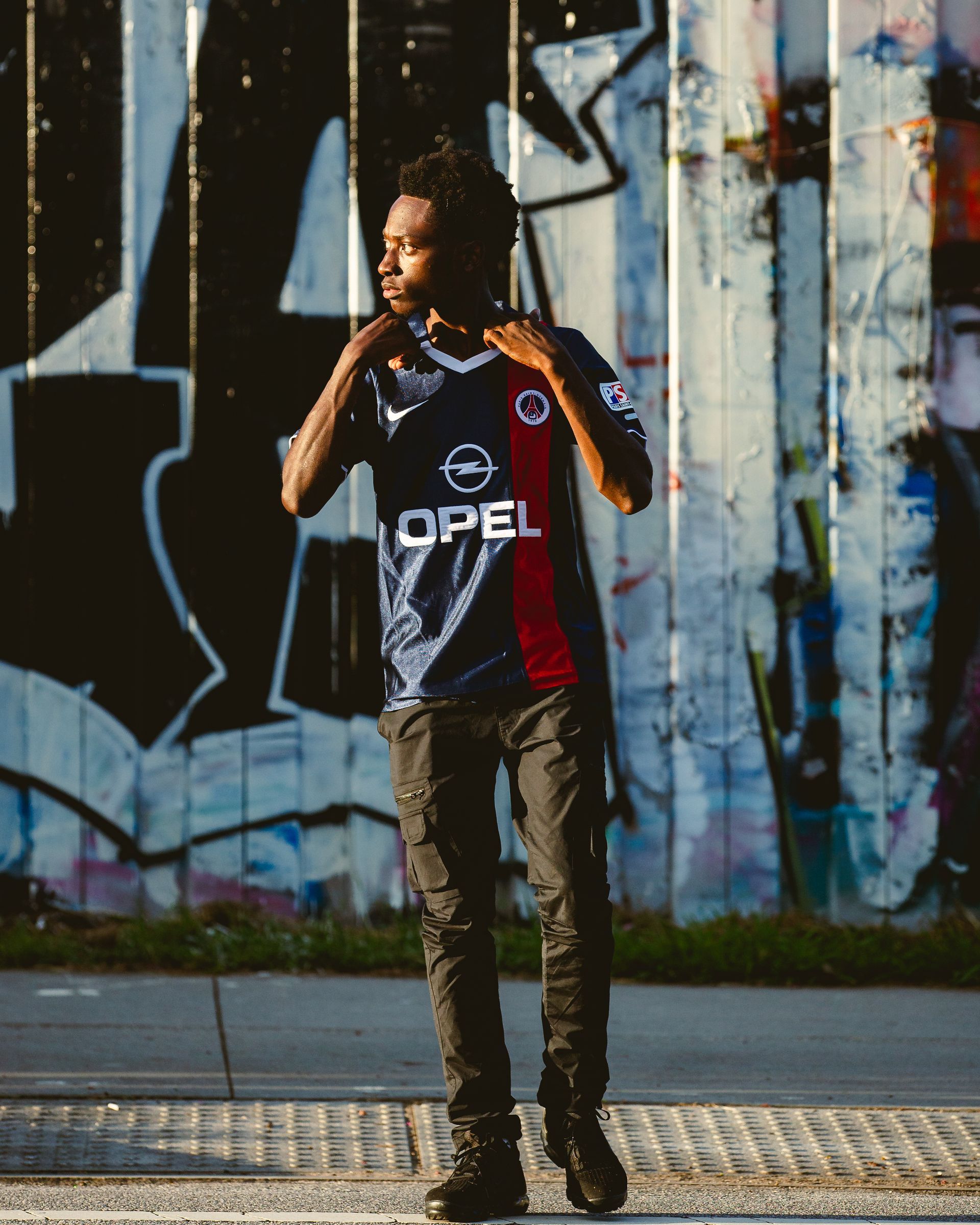 Man in a soccer jersey stands in front of a graffiti-covered wall, adjusting his collar.