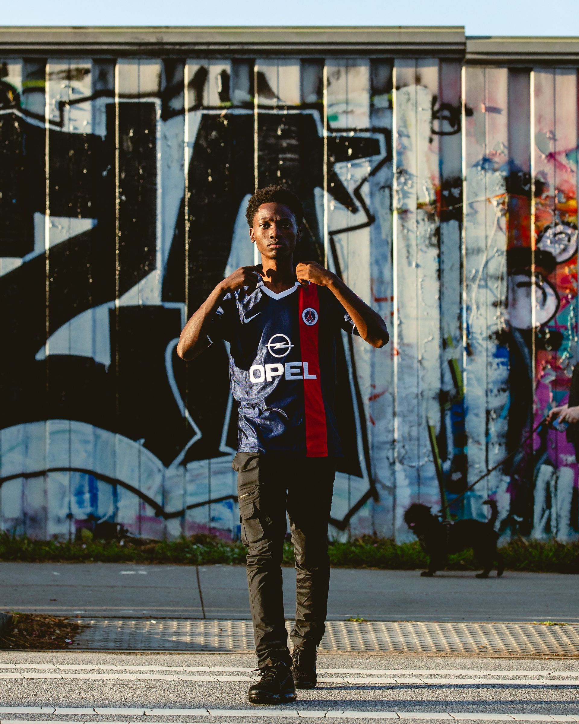 Man in a PSG jersey and black pants stands in front of a graffiti-covered wall, holding jersey up.