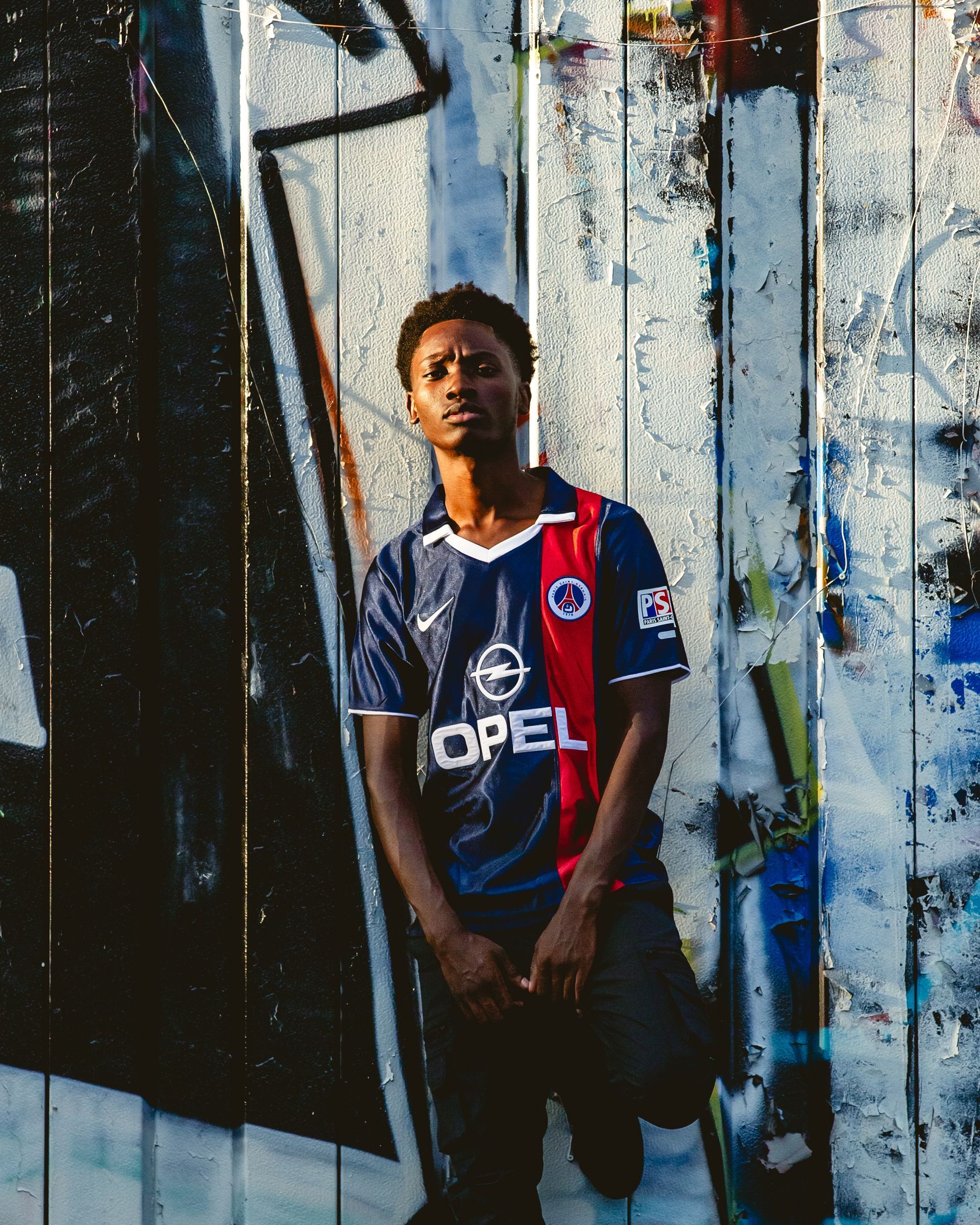 Young person leans against a graffiti-covered wall, wearing a blue and red soccer jersey.