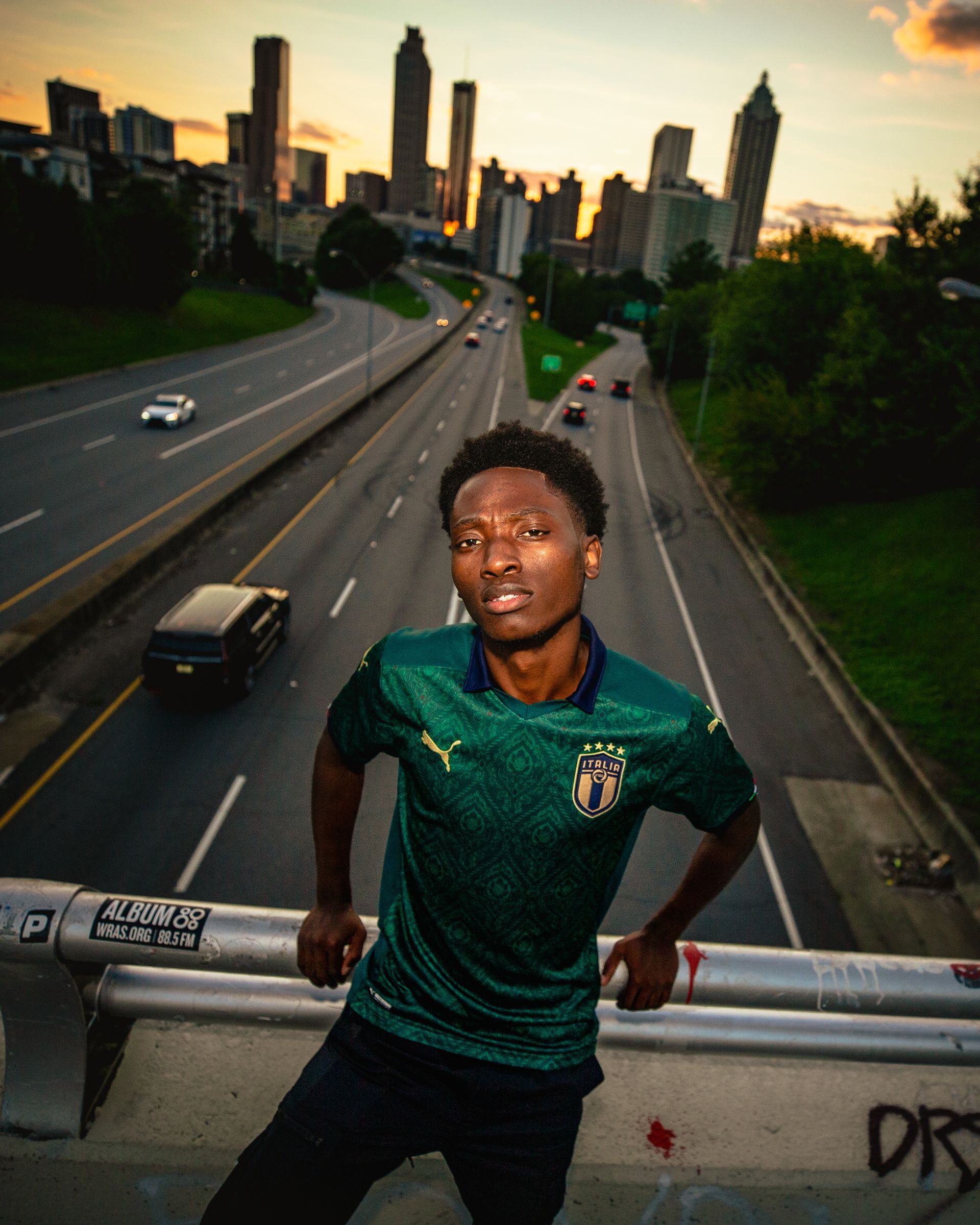 Man in green jersey leans on a bridge railing, Atlanta skyline in background; highway traffic.