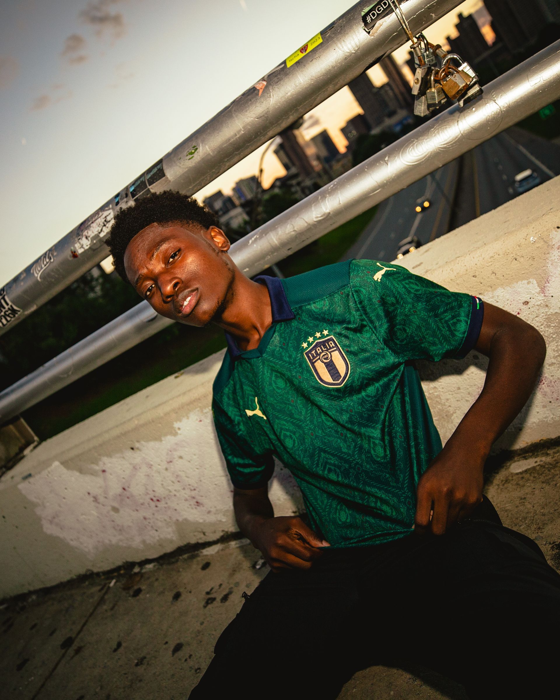 Person wearing a green soccer jersey, leaning against a railing overlooking a city at dusk.