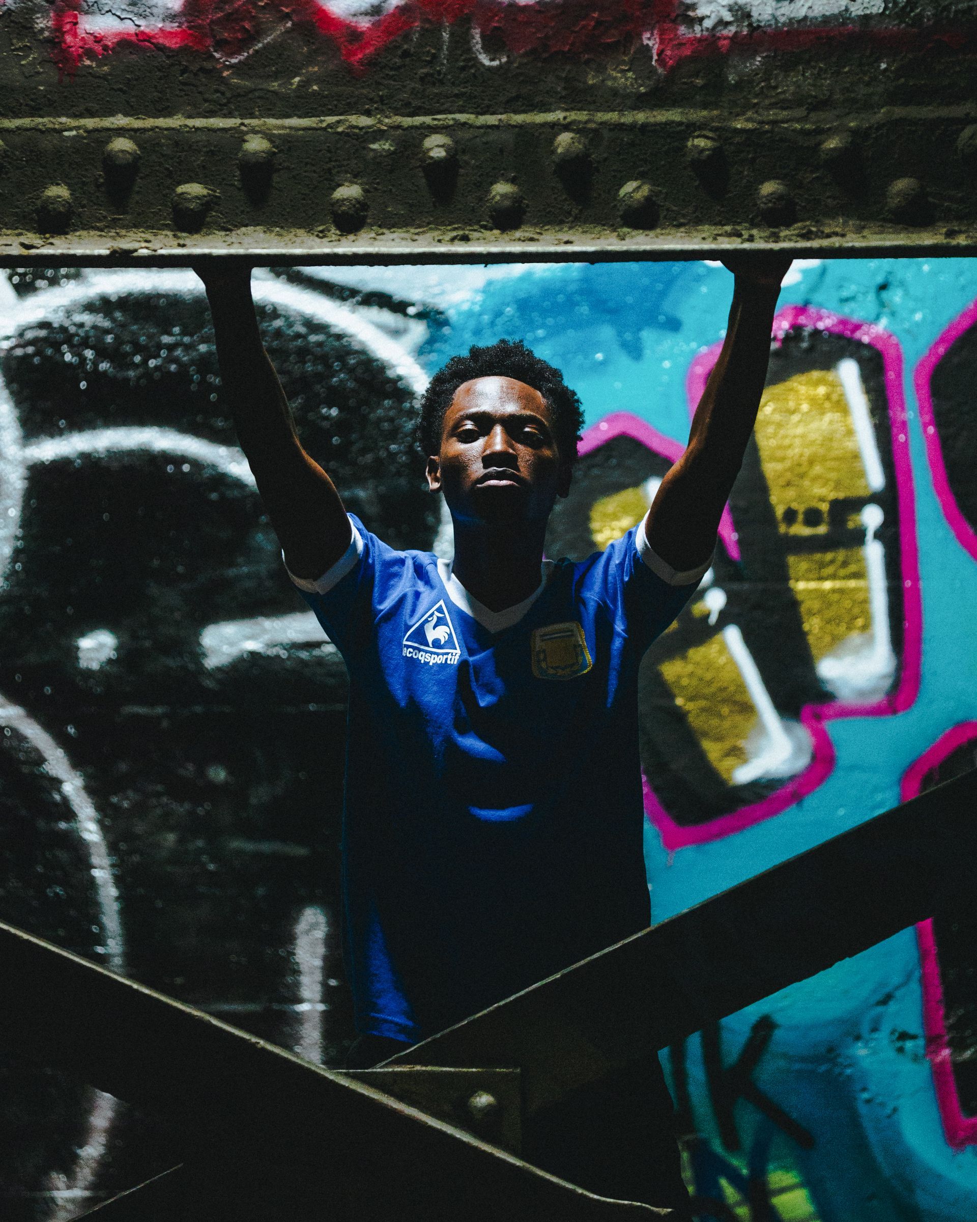 Person in blue soccer jersey raises arms, standing under metal beams, in front of graffiti.