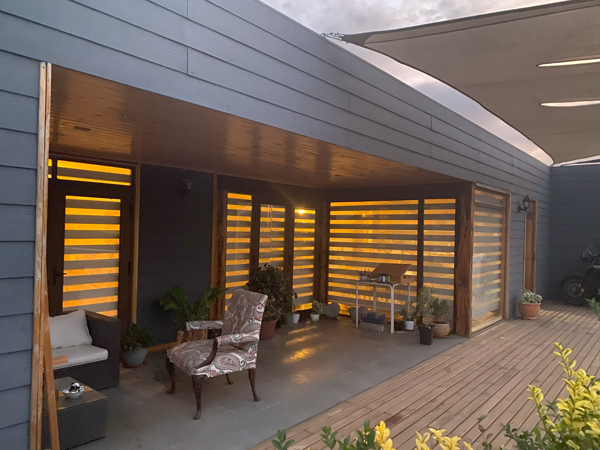 Patio con terraza de madera, revestimiento gris y persianas, iluminado desde el interior. Muebles y plantas visibles.