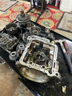 Disassembled car engine parts on a black workbench with tools and a patterned rug nearby