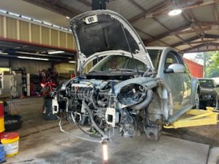 Damaged silver car in a workshop, hood open and front end stripped, showing exposed engine components.