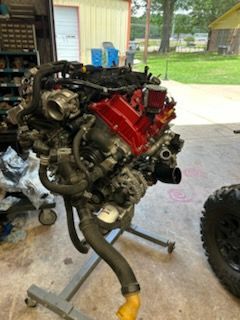 Partially assembled V8 engine on a stand in a garage, with red valve covers and exhaust headers visible.