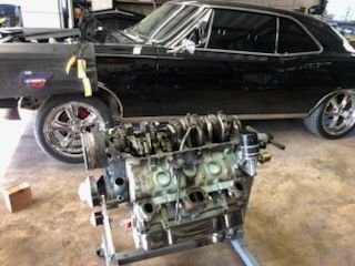 Car engine on a stand in a garage, with a black car in the background.
