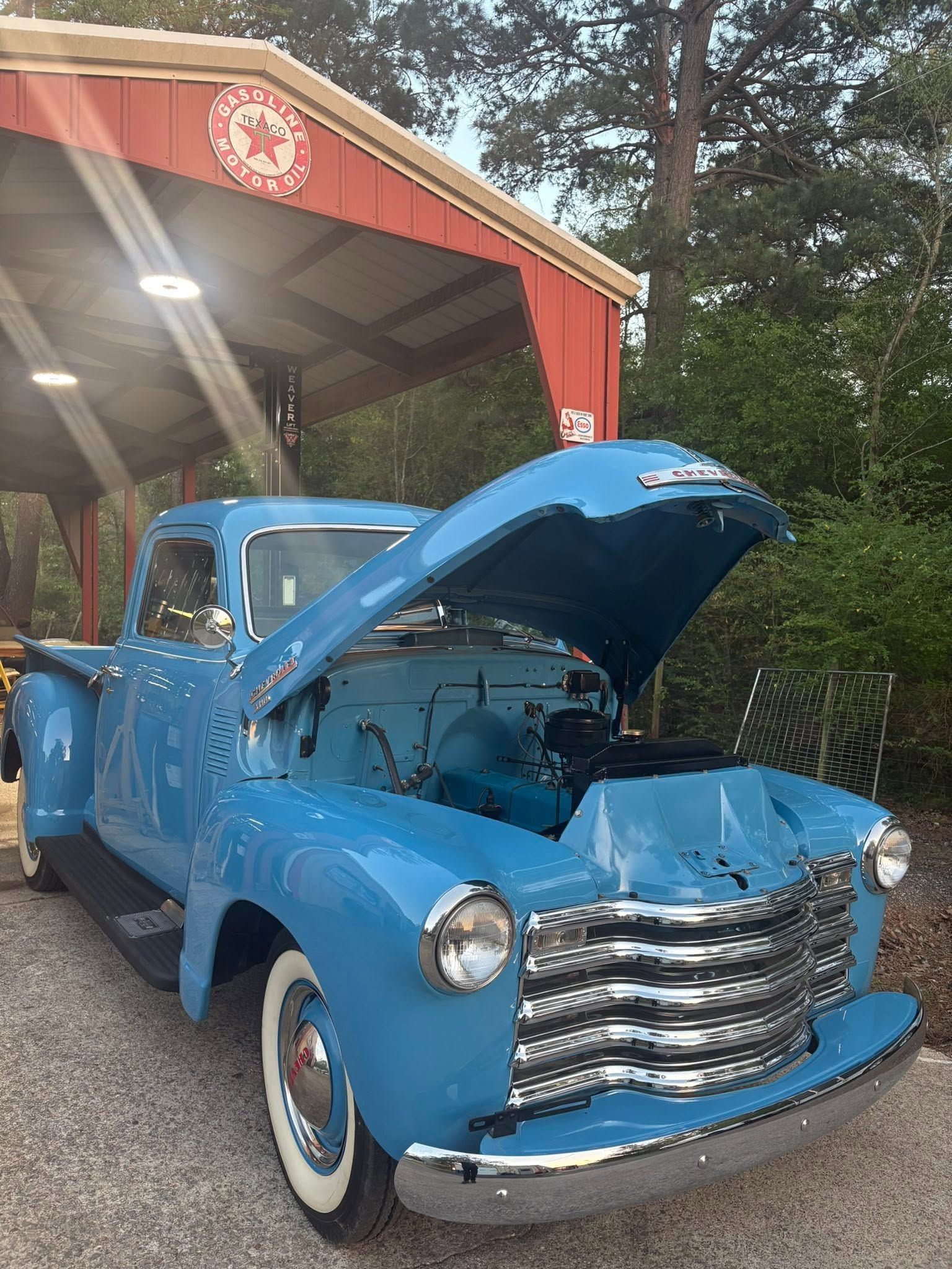 Blue vintage pickup truck with hood open, parked under a red-roofed shelter outdoors
