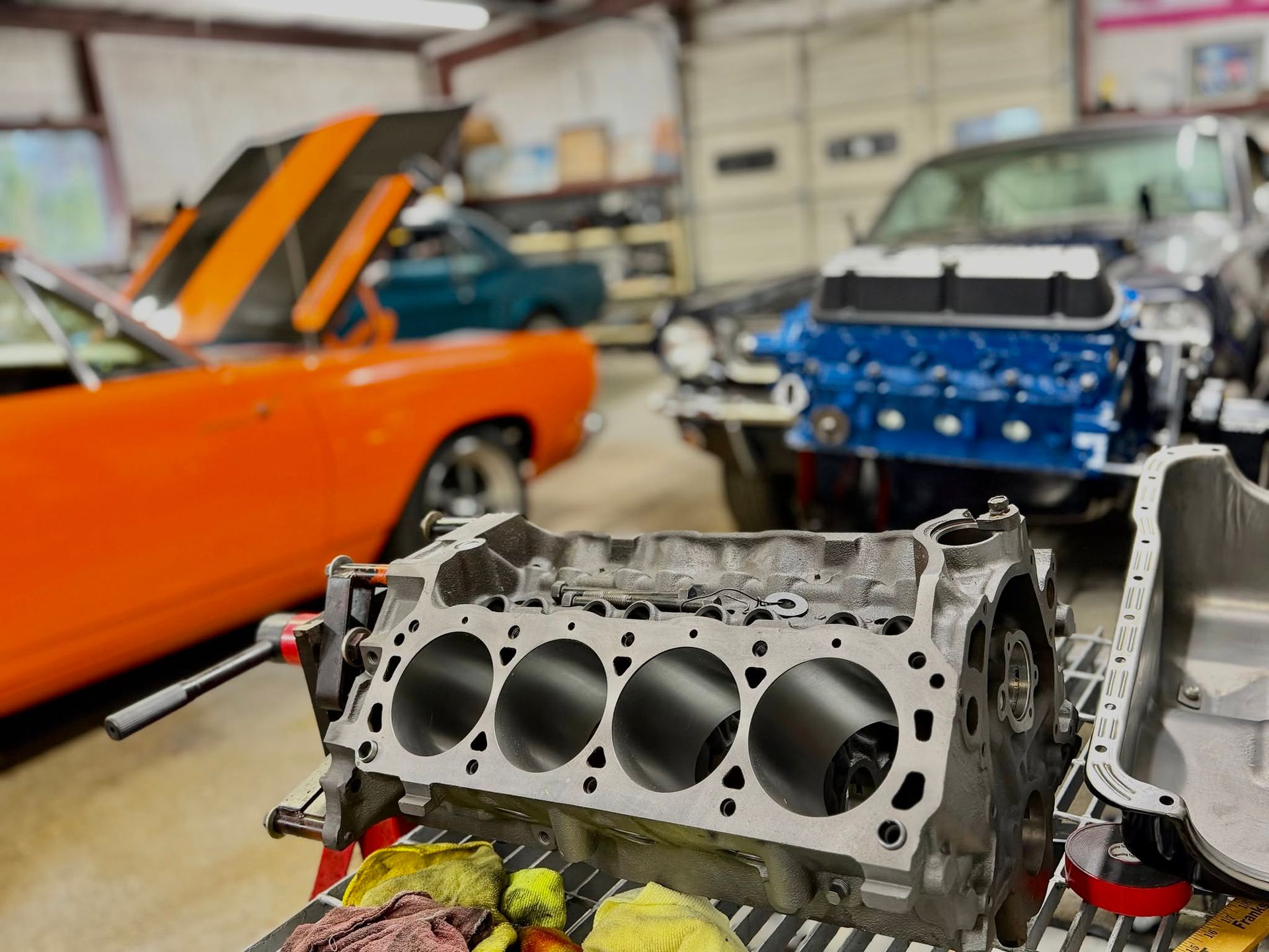 Engine block in workshop with orange car, blue car, and a man working in the background