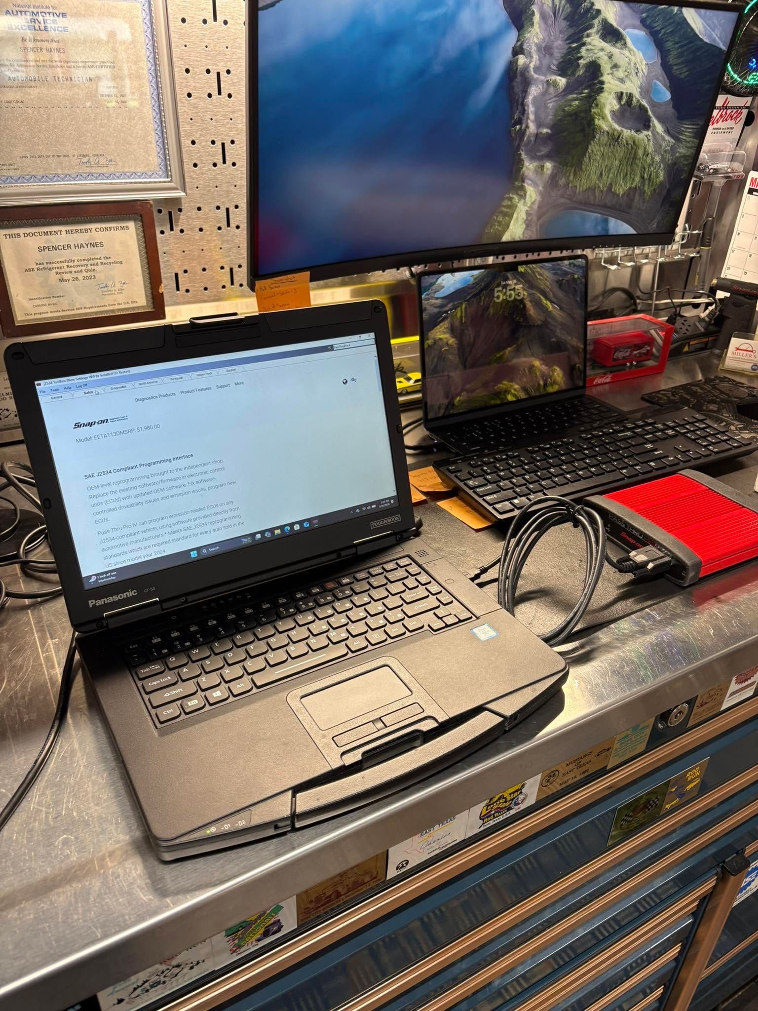 Two laptops on a workbench, one open to a text document and one beside a large monitor in a workshop.