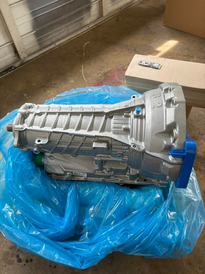 Silver car transmission resting on a blue plastic tarp outdoors, with a cardboard box nearby.
