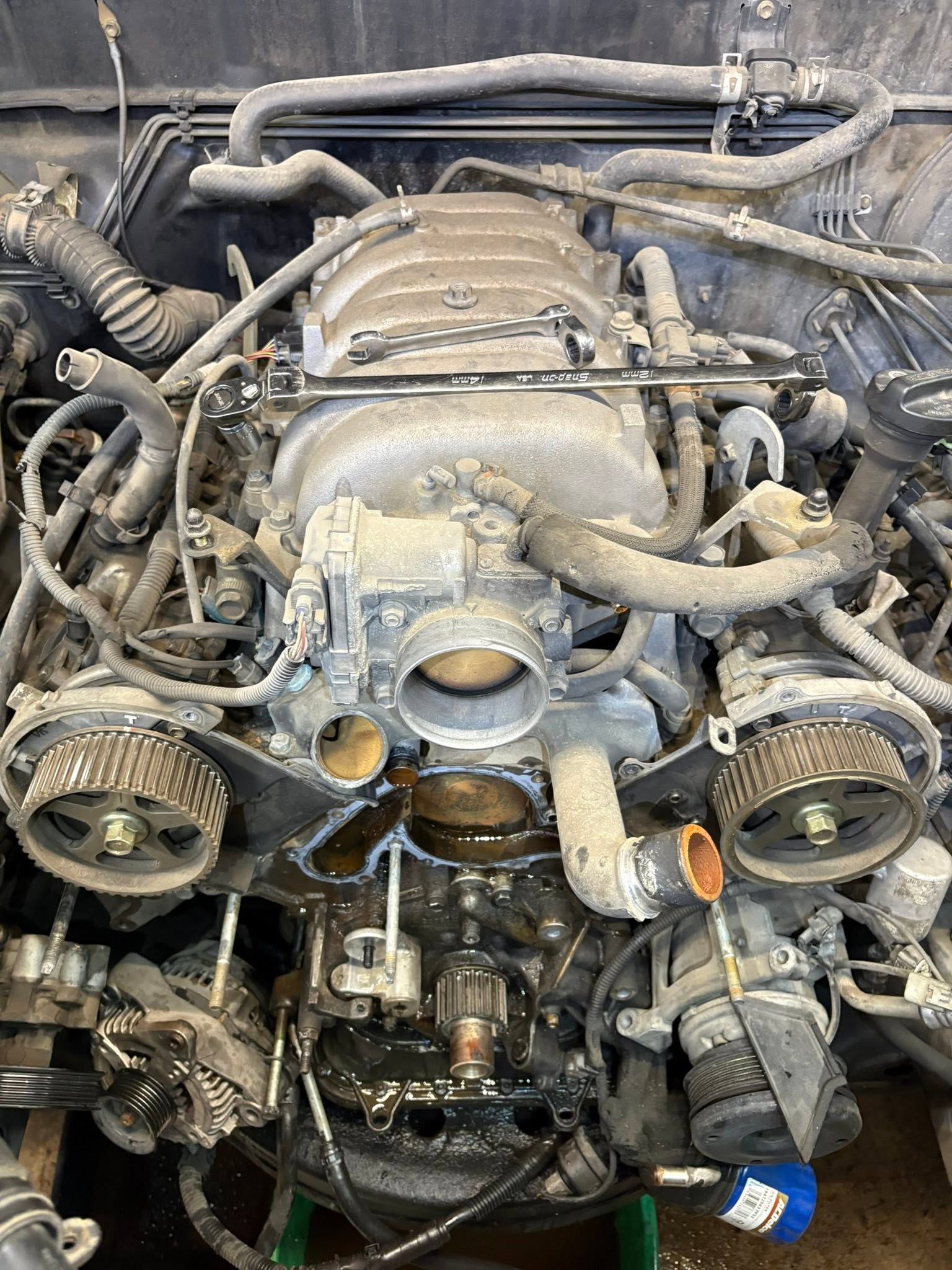 Exposed car engine bay with hoses, belts, and intake manifold, viewed from above.